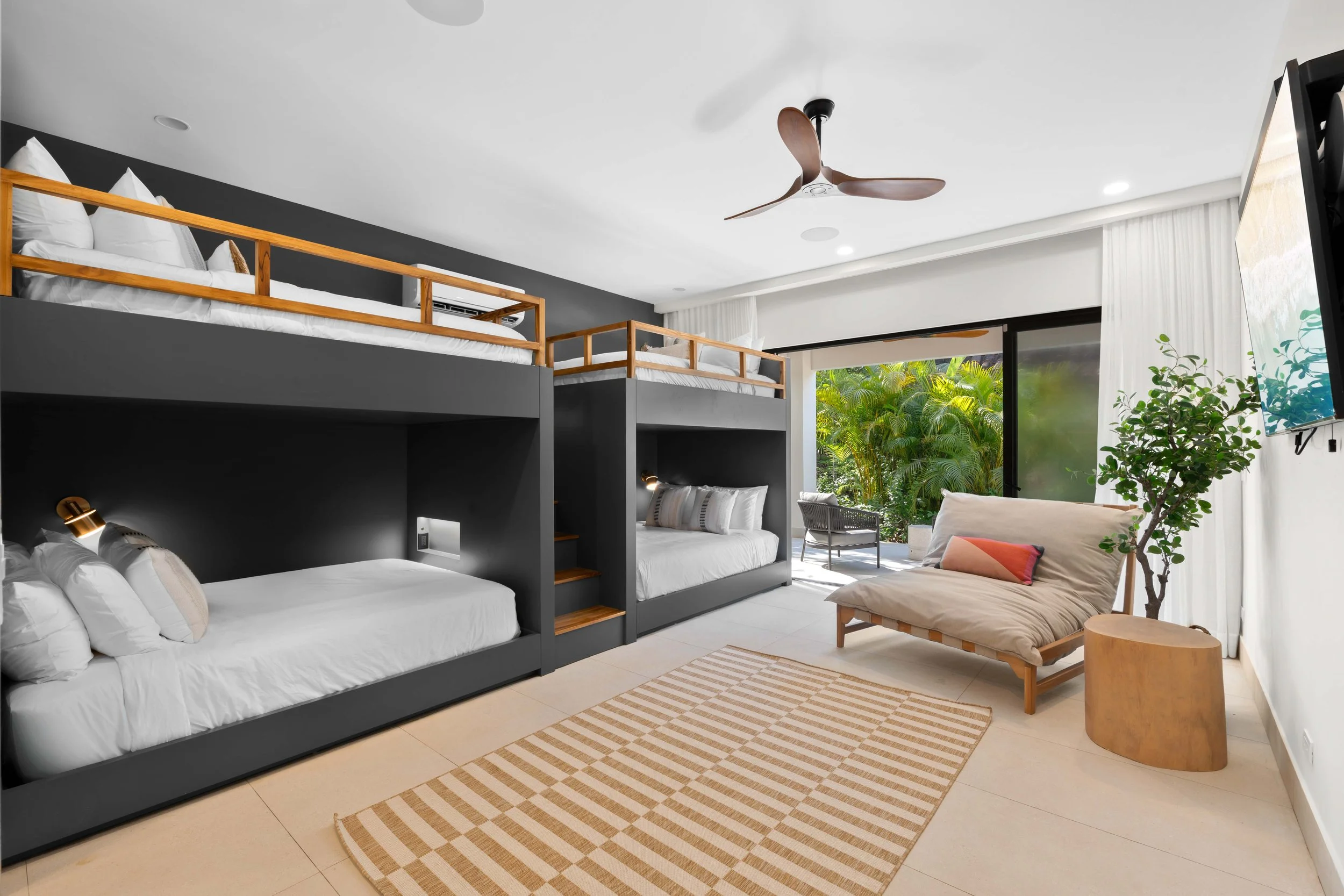 Modern hotel room with two sets of bunk beds, one with a dark frame and bedding, and one with a dark frame and white bedding. There is a lounging chair, a small wooden side table, a large window with curtains, and a view of green foliage outside. A c