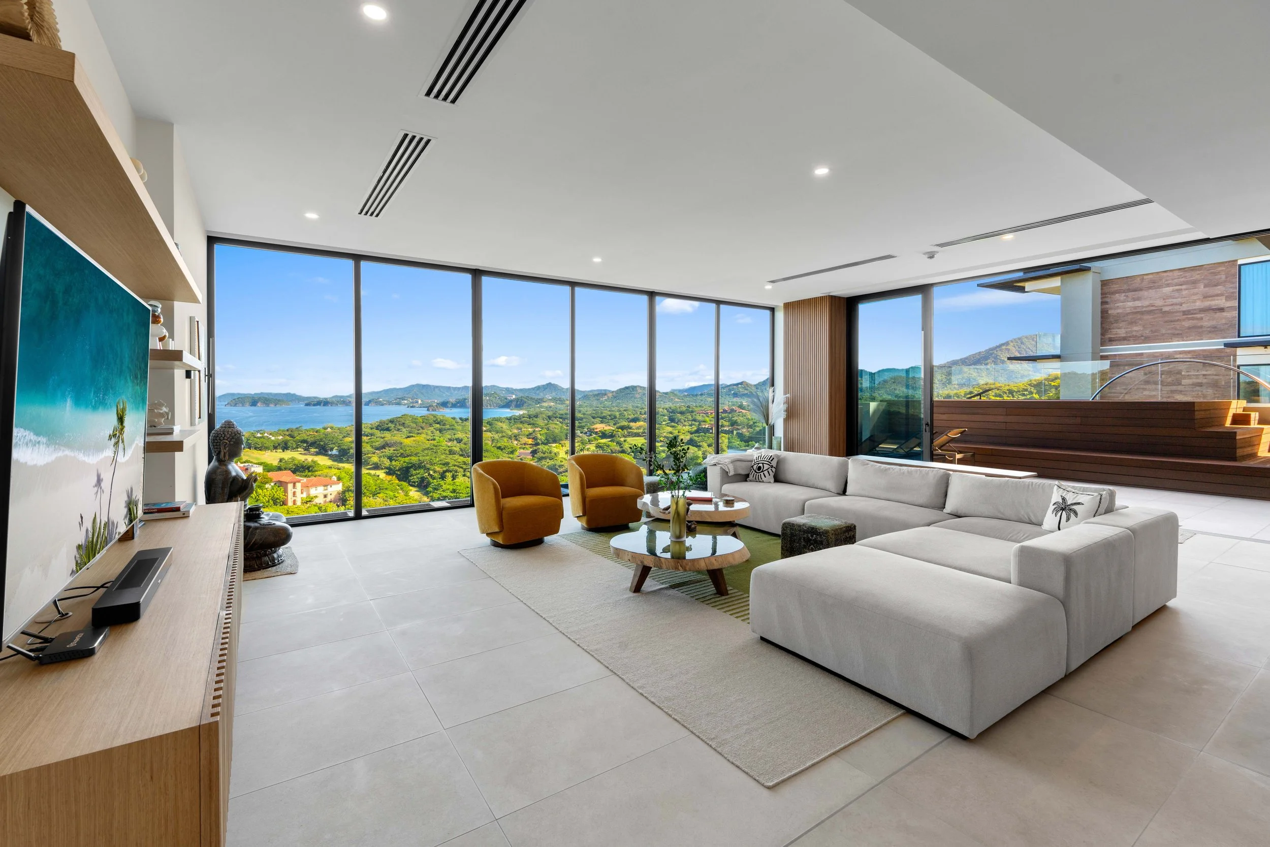 Modern living room with large floor-to-ceiling windows offering a scenic view of a lush green landscape and distant mountains, furnished with a white sectional sofa, two mustard-colored armchairs, a wooden coffee table, and decorative items.