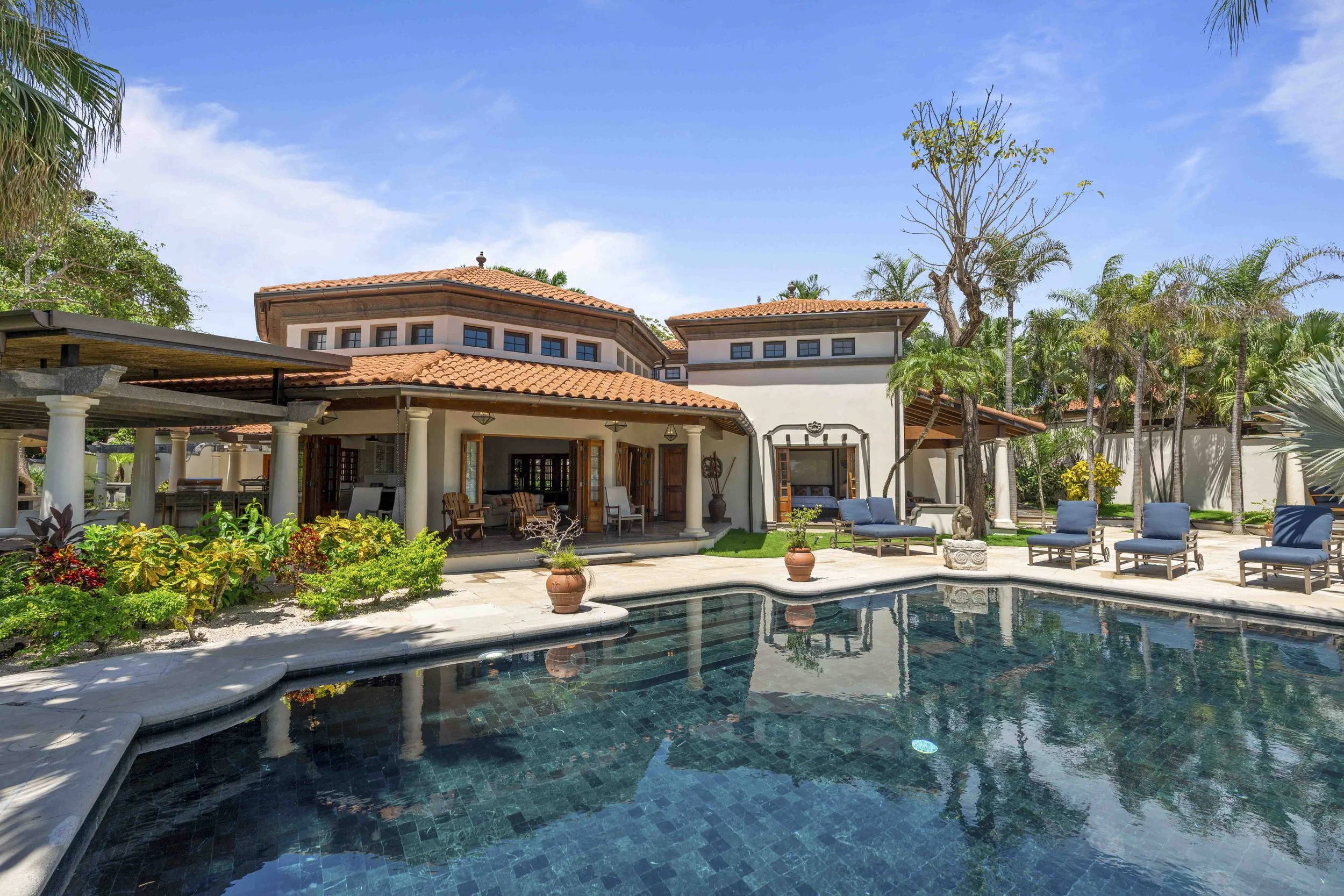 Luxury house in Tamarindo, Costa Rica with a swimming pool in the backyard, outdoor seating, lush green trees, and a blue sky.