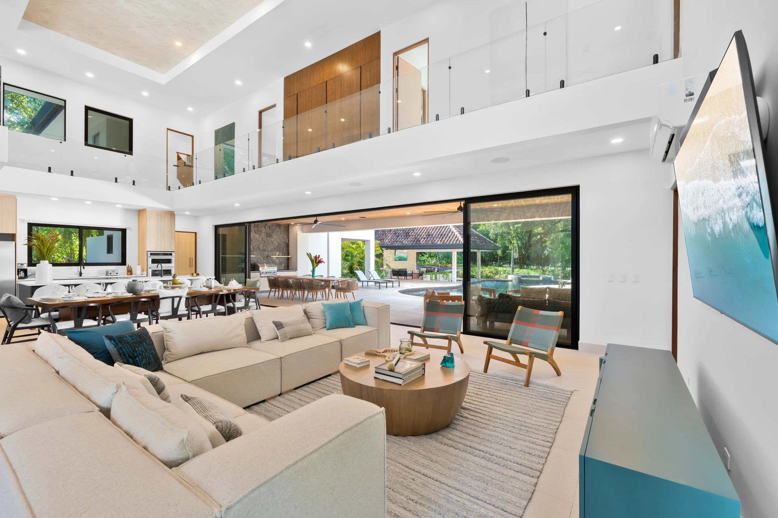 Modern open-concept living room and kitchen with large windows, white walls, beige sectional sofa, colorful chairs, wooden dining table, outdoor patio, and swimming pool in the backyard.
