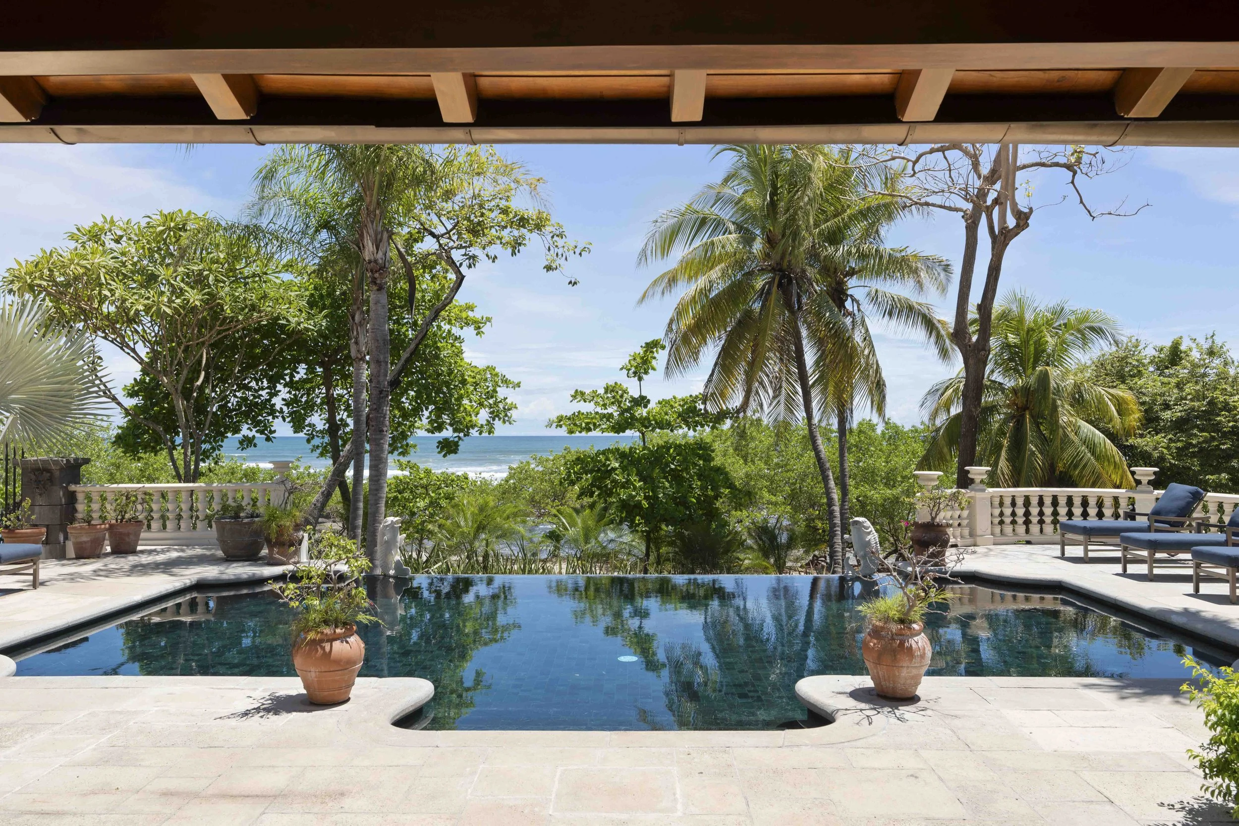 Poolside view in Tamarindo, Costa Rica with outdoor seating, potted plants, and lush green trees including palm trees, overlooking the ocean on a sunny day.