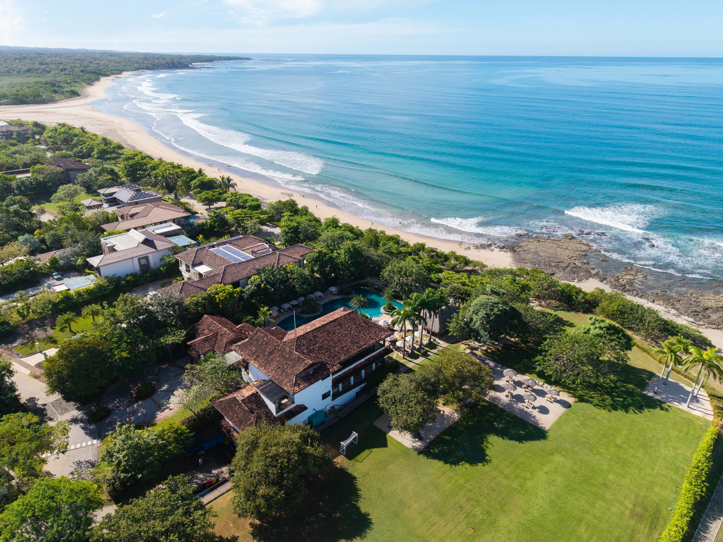An aerial view of a coastal area featuring a beach, ocean waves, lush greenery, and residential houses with a swimming pool.