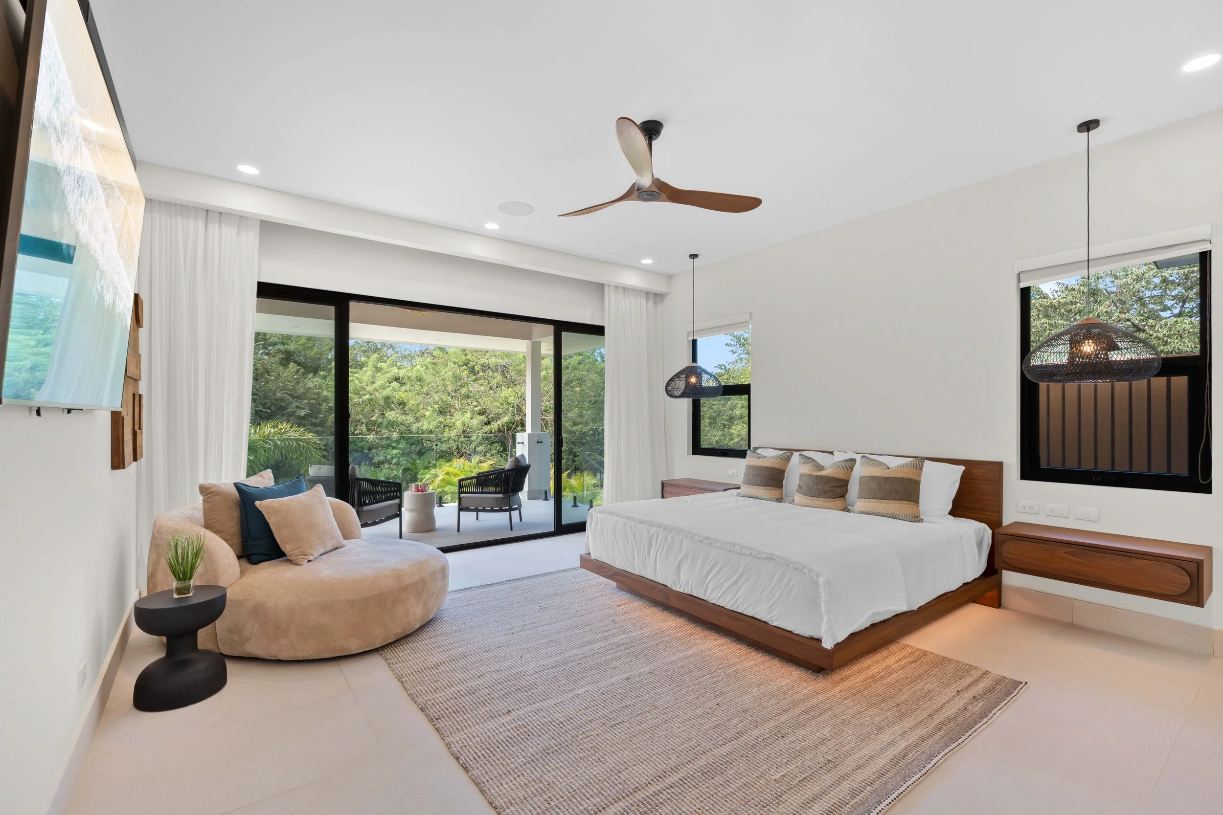 Bedroom with a large bed, bedside tables, a beige plush chair with pillows, sliding glass door leading to a balcony with outdoor seating, white walls, black window frames, ceiling fan, modern lighting, and a rug on the floor.