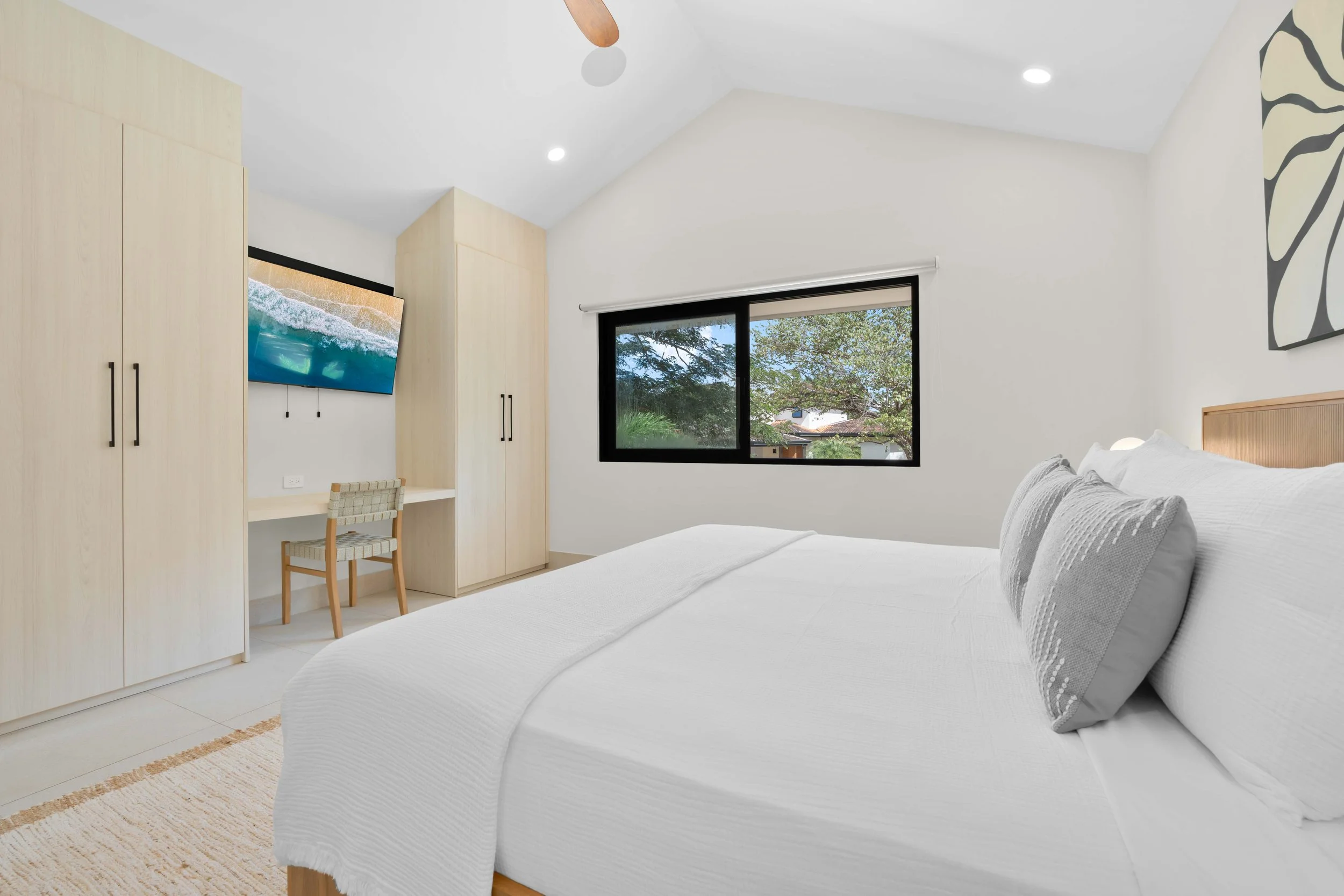 Clean, bright bedroom with a large bed, white bedding, gray pillows, a window showing green trees outside, and a built-in wardrobe, with a wall-mounted TV and a small desk with a chair.