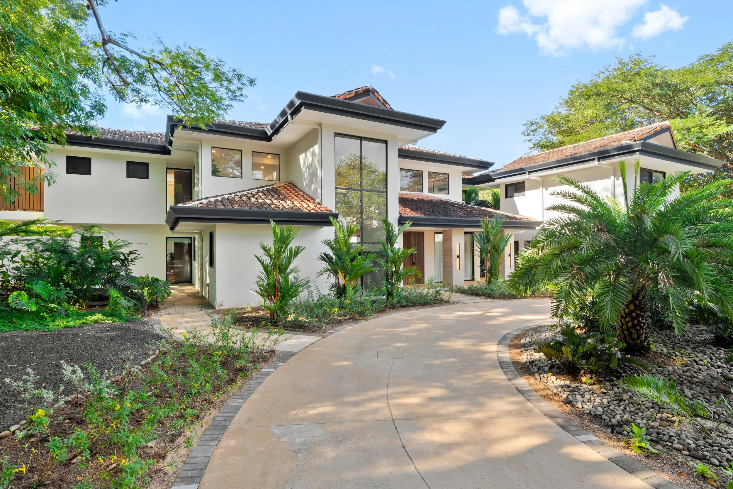 Modern two-story house with white walls, large glass windows, and a tiled roof, surrounded by lush greenery and palm trees, with a curved driveway in front.
