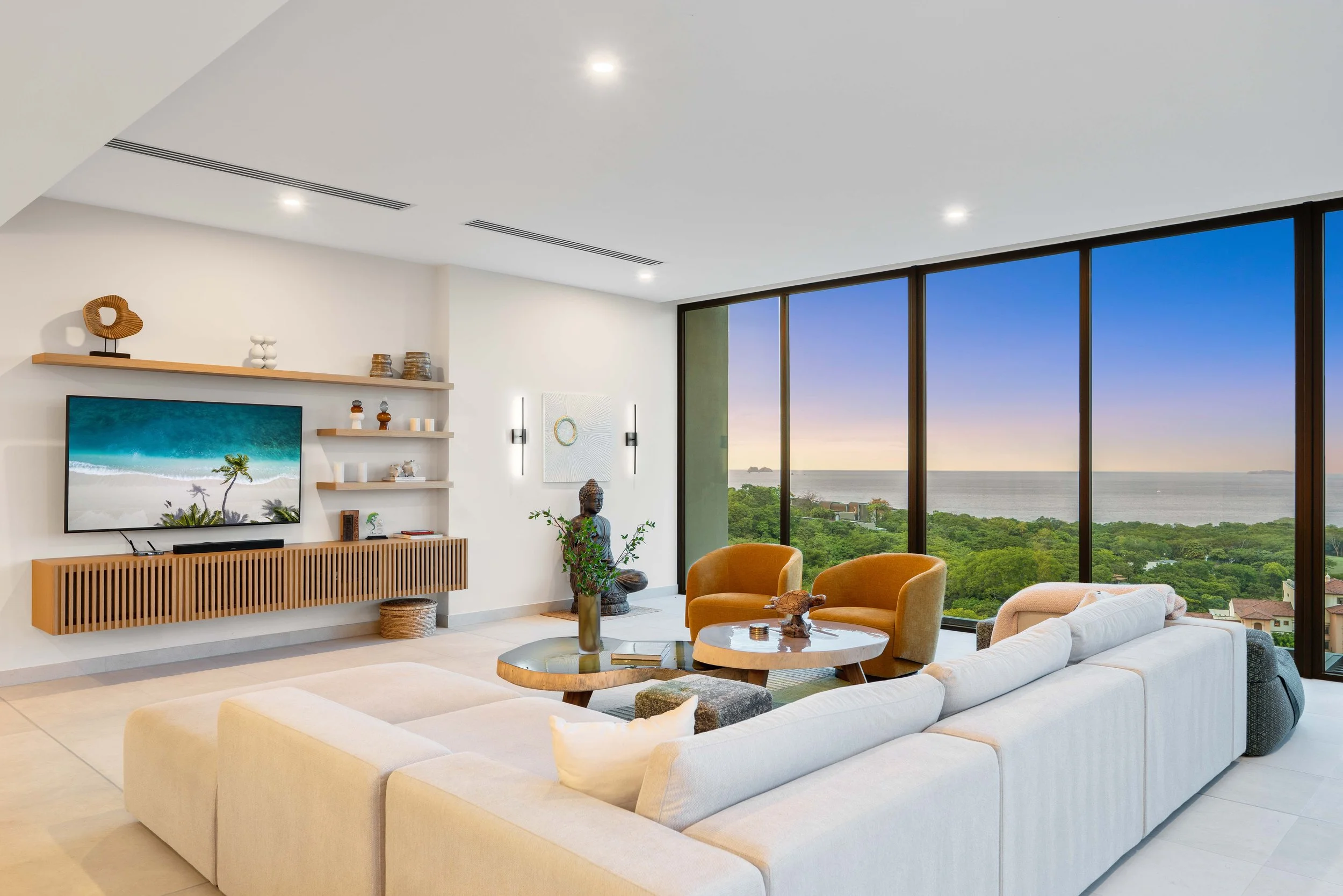 Modern living room with large floor-to-ceiling windows overlooking a scenic view of greenery and water at sunset, white sectional sofa, round coffee tables, two mustard-colored armchairs, decorative plants, art pieces, and a television mounted on the