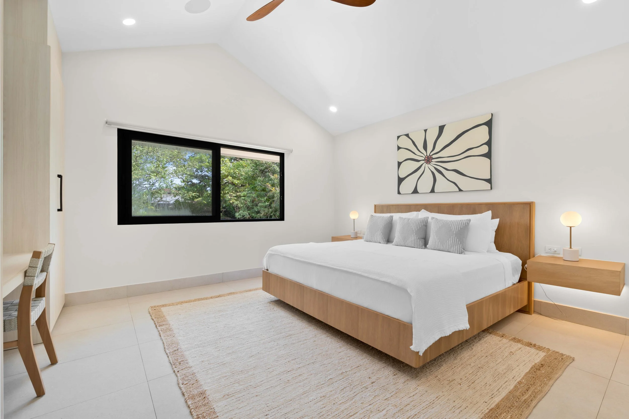 Modern bedroom with a large bed, white bedding, gray pillows, wooden headboard, bedside tables with lamps, a window showing green trees, and abstract wall art.
