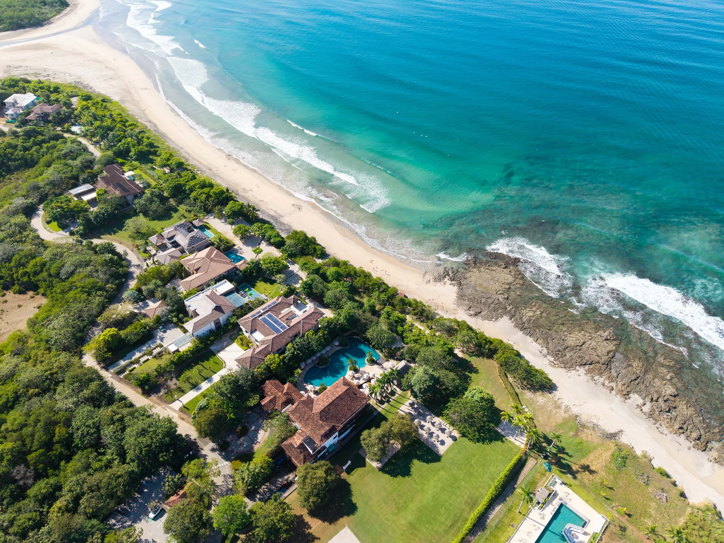 Aerial view of a beachfront residential neighborhood with houses, trees, and a sandy beach with turquoise ocean waves.