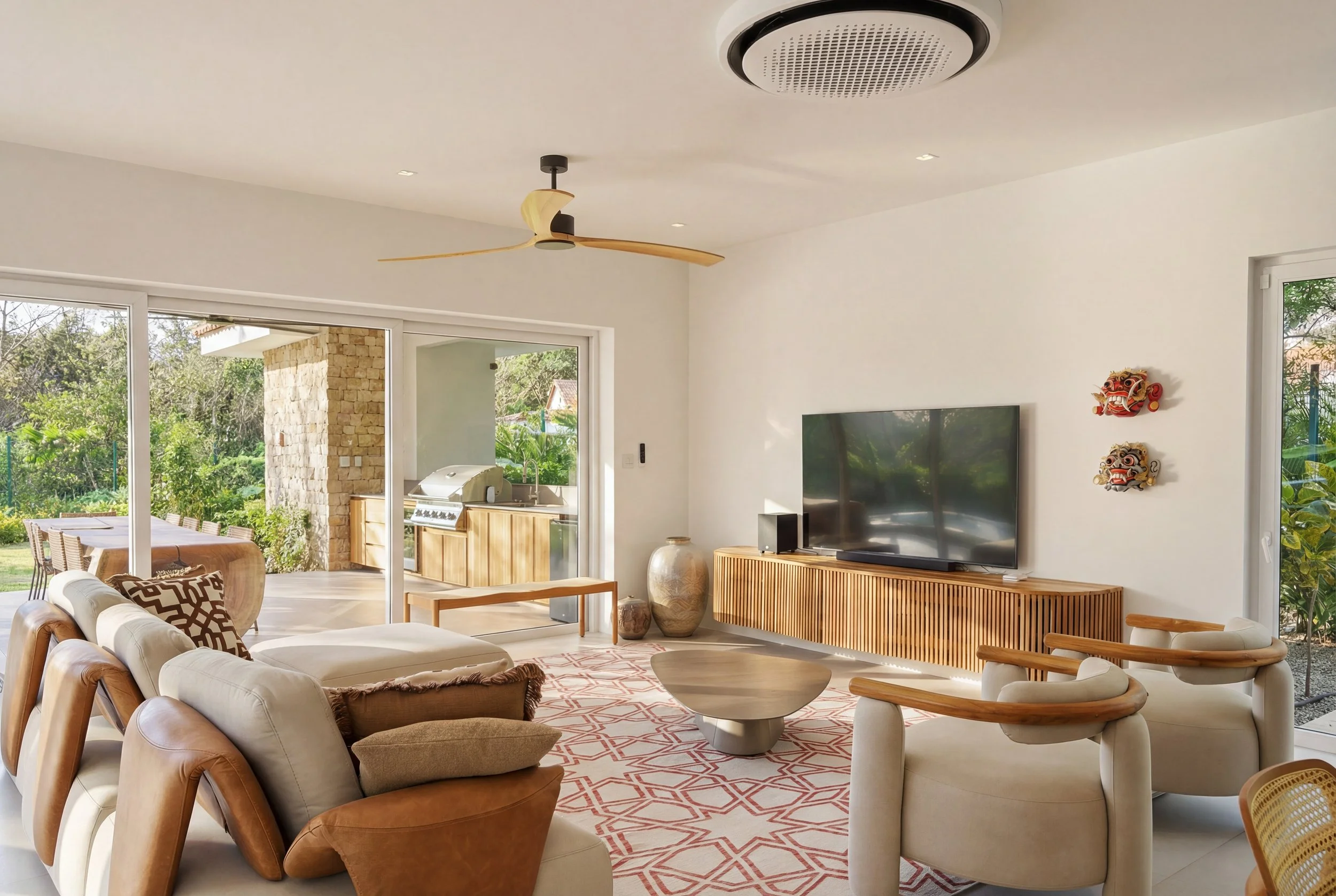 Modern living room with white walls, sliding glass doors leading to outdoor patio, wooden furniture, flat-screen TV on a slatted wooden console, two decorative masks on the wall, ceiling fan, patterned rug, and outdoor barbecue area visible through t
