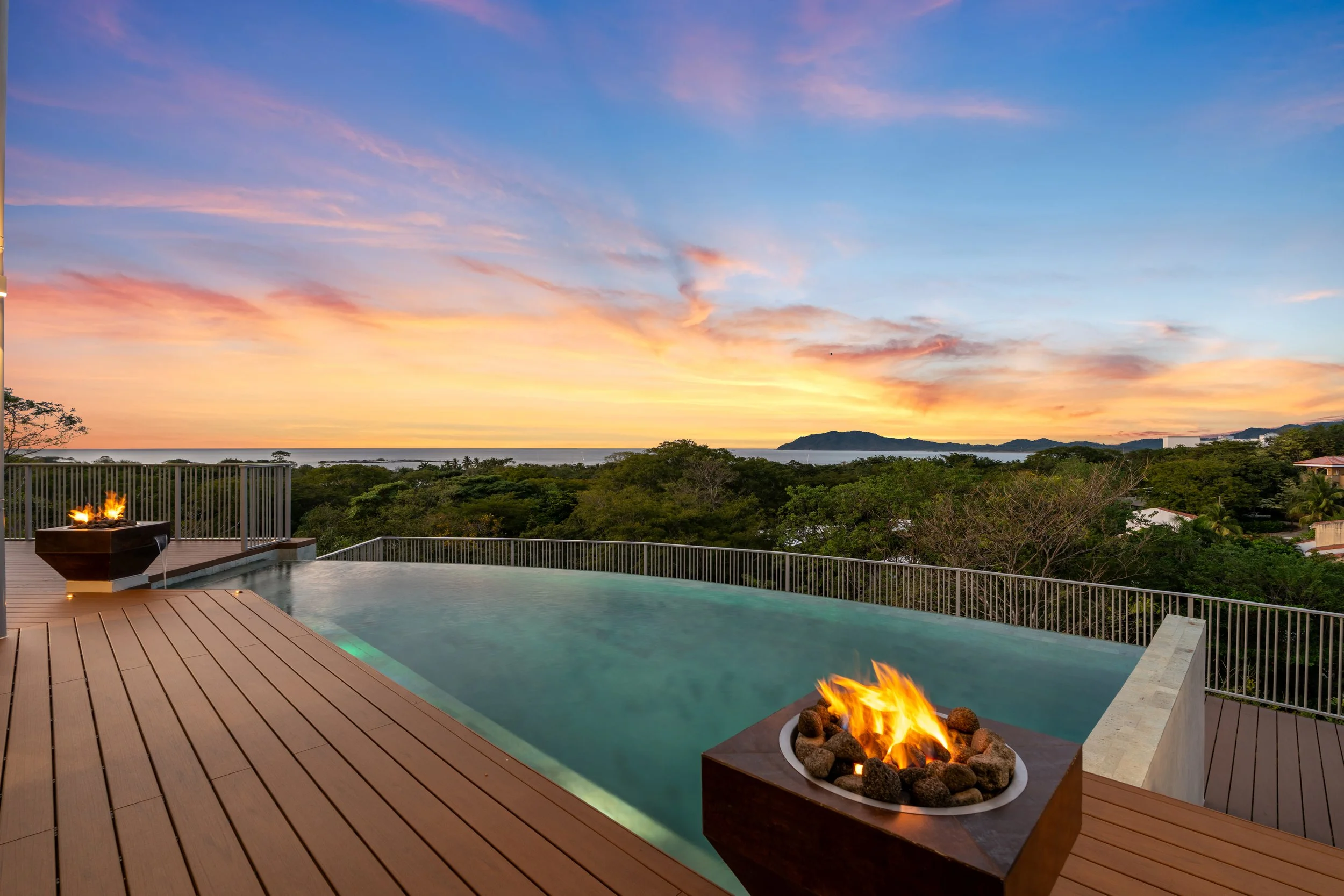 Infinity pool in Tamarindo, Costa Rica with fire features on a wooden deck overlooking trees, hills, and the ocean at sunset.
