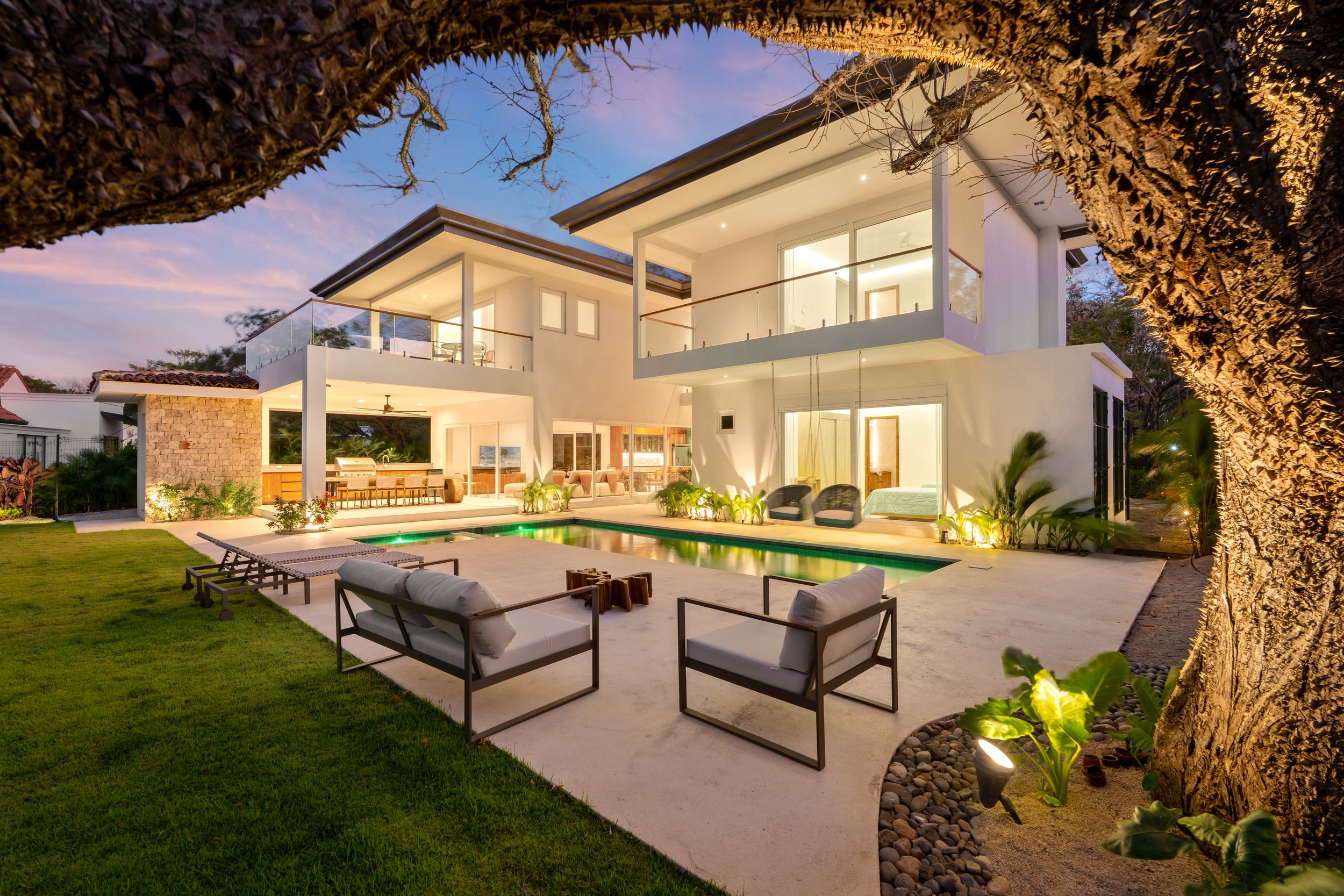 Modern two-story house with large windows and balconies, illuminated outdoor seating area, swimming pool, lawn, and garden with trees and plants at dusk.