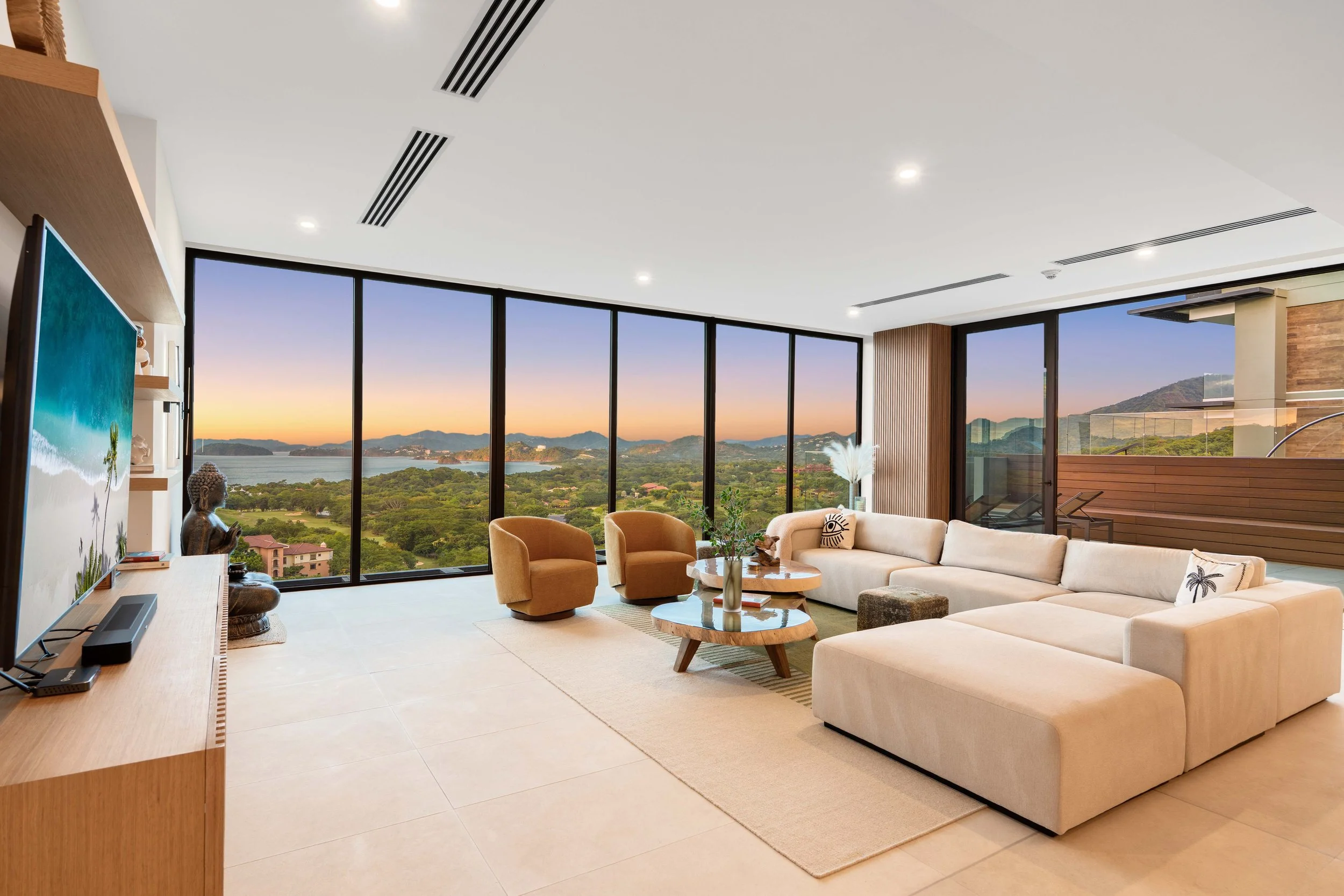 Modern living room with large windows overlooking a scenic landscape during sunset, with a beige sectional sofa, two tan armchairs, a wooden coffee table, plants, and a flat-screen TV.