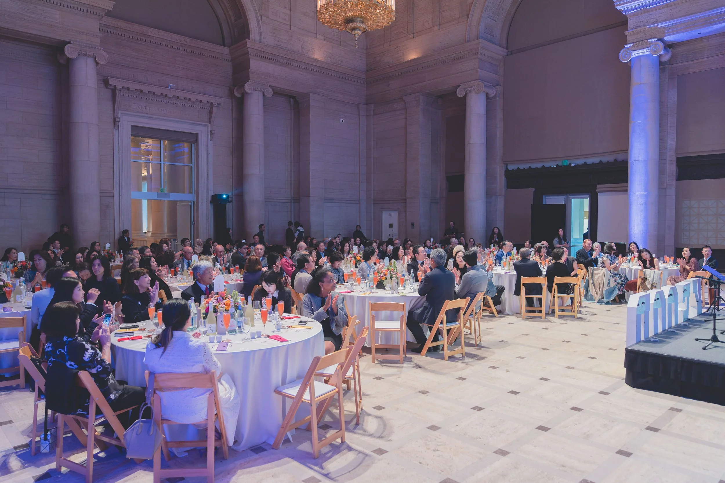 A large formal event or banquet taking place in an elegant, grand hall with high ceilings, large columns, and ornate lighting fixtures. Attendees are seated at round tables with white tablecloths, decorated with floral centerpieces and orange-colored