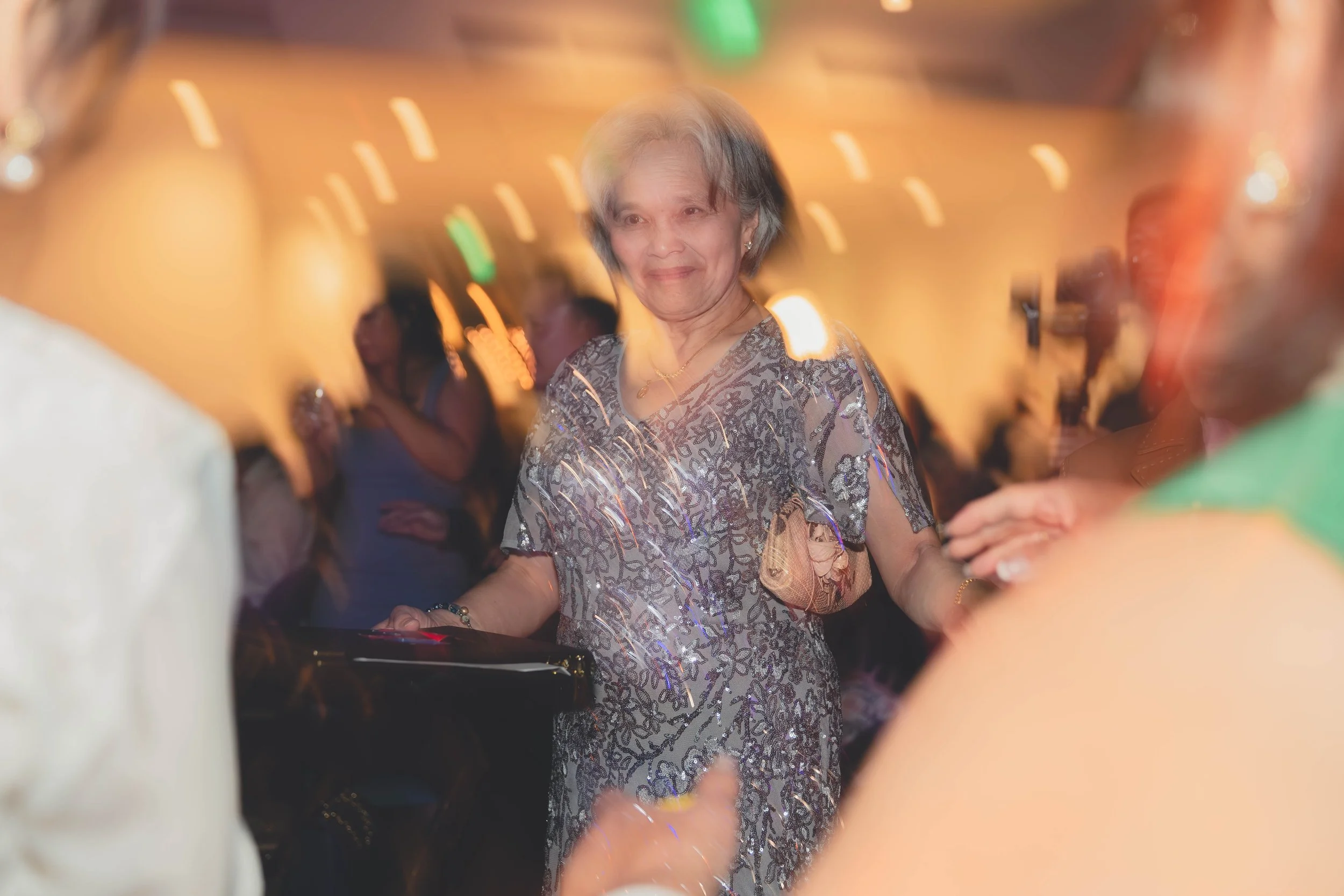 Blurred image of a smiling elderly woman in a sparkly dress at a party, surrounded by people and lights.