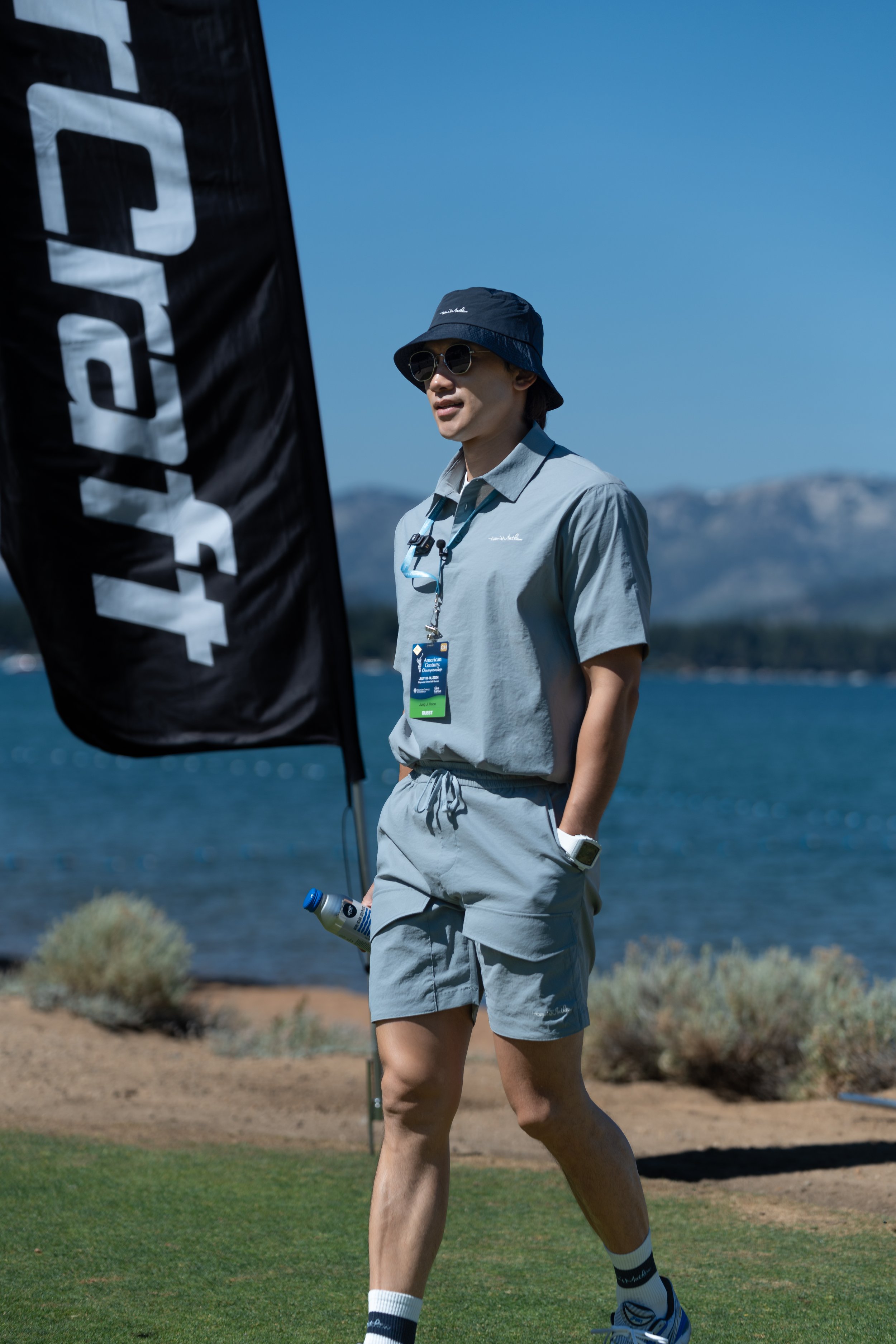 Person in casual clothing with bucket hat and sunglasses walking near a lake.