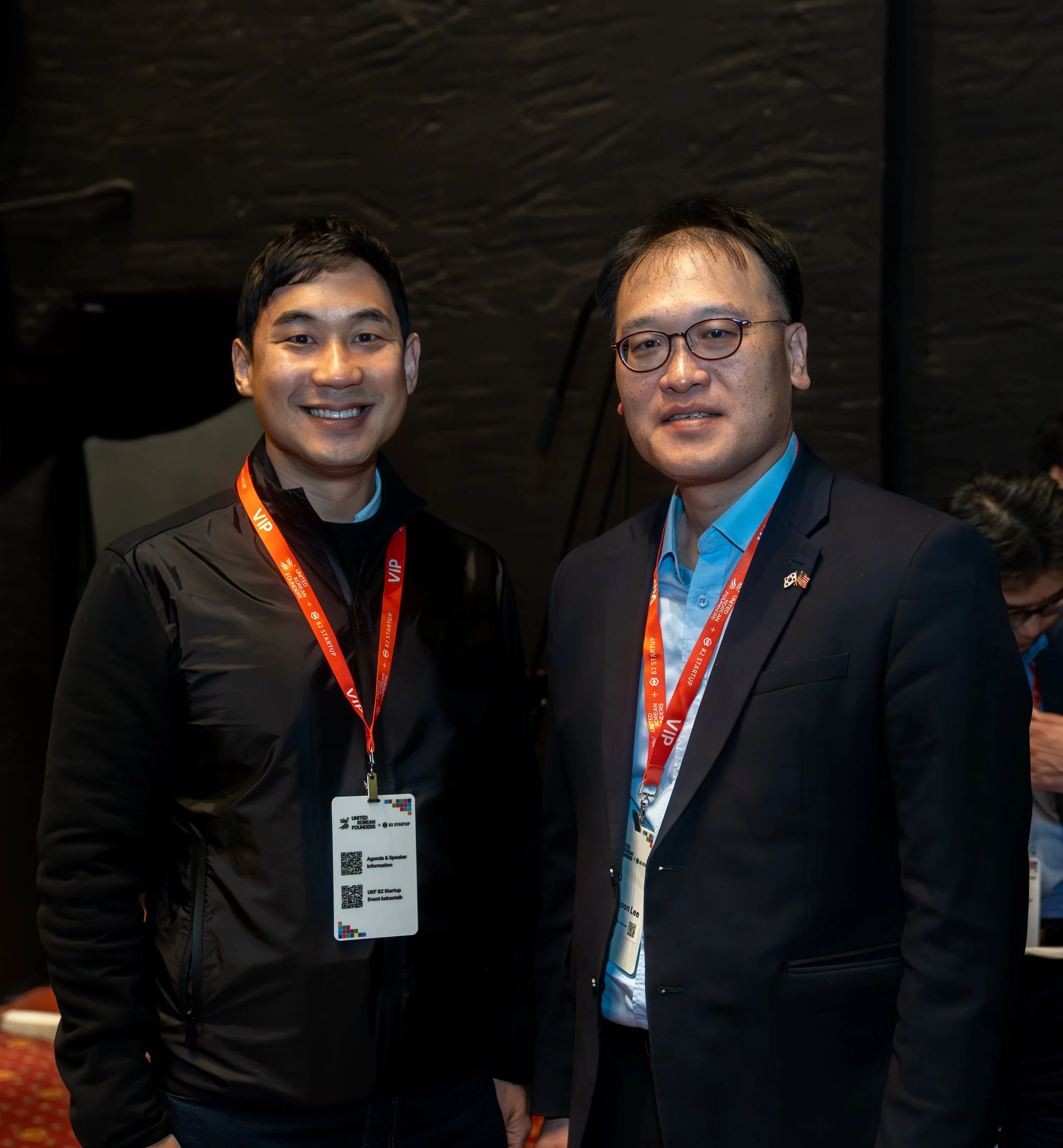 Two men wearing conference badges and red lanyards, standing indoors, dressed in business attire.