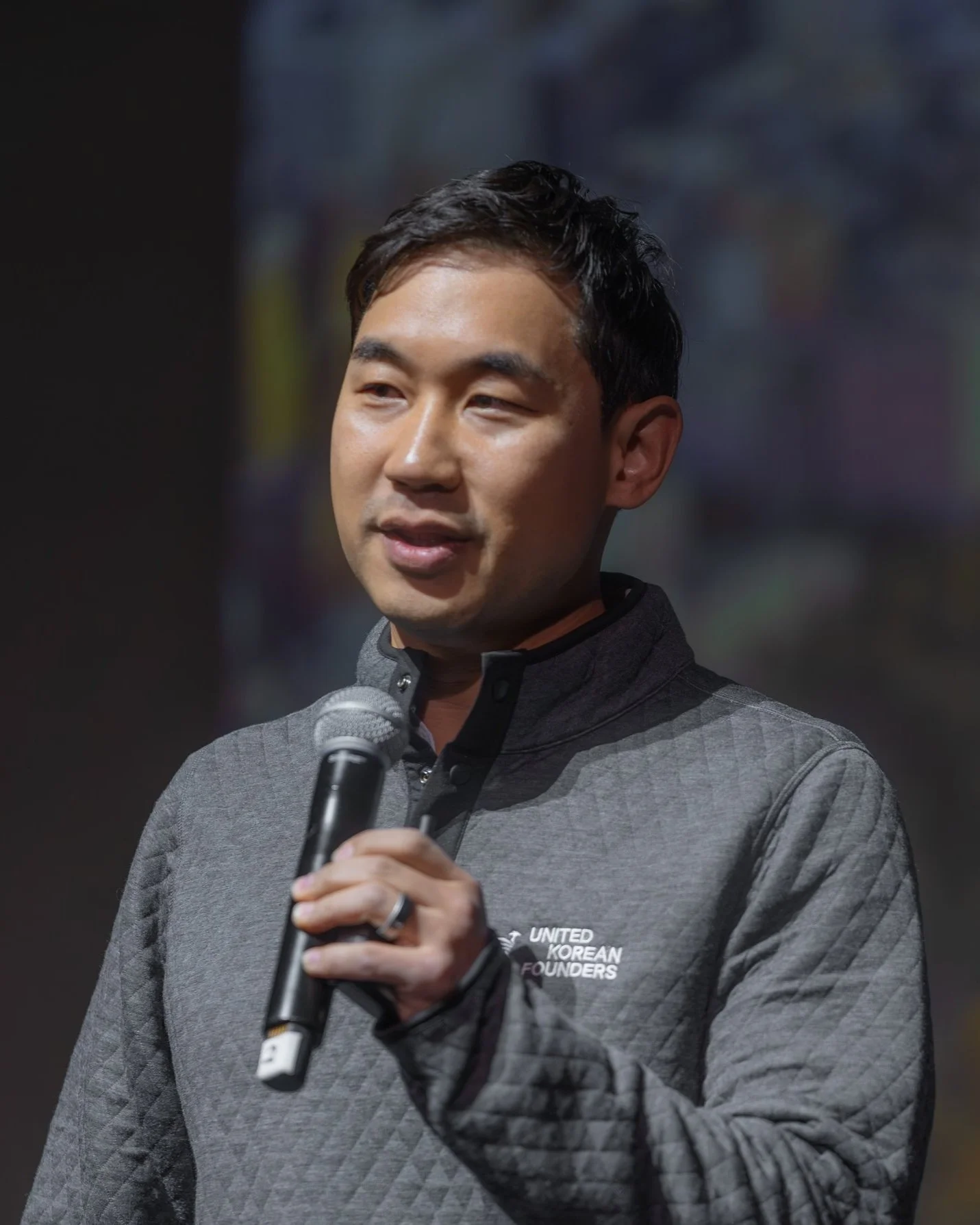 Person speaking into a microphone, wearing a gray quilted jacket with 'United Korean Founders' text.