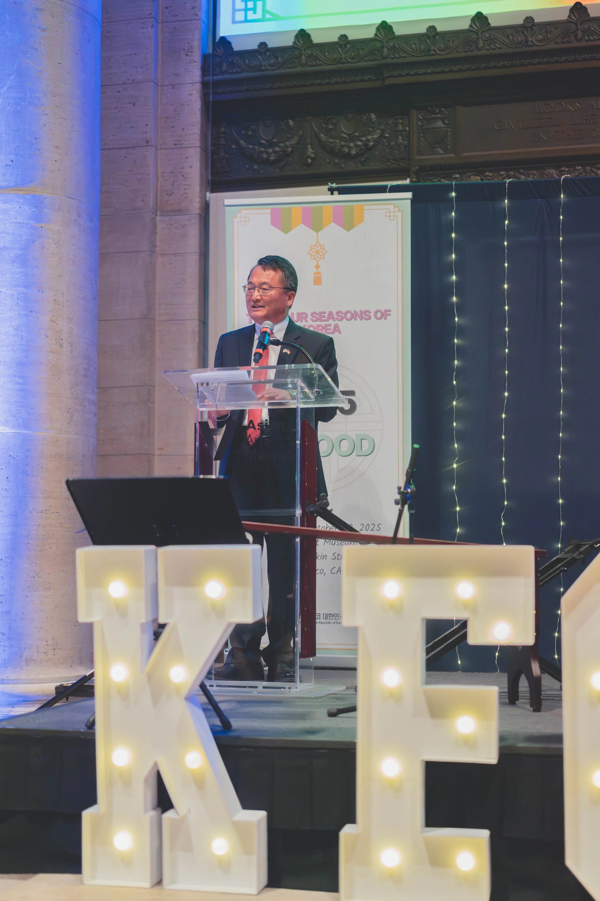 A man in a suit and red tie is speaking at a podium during an event, with large illuminated letters 'K' and 'E' in front of the stage. There is a backdrop with a poster and string lights behind him, in an indoor setting with stone columns.