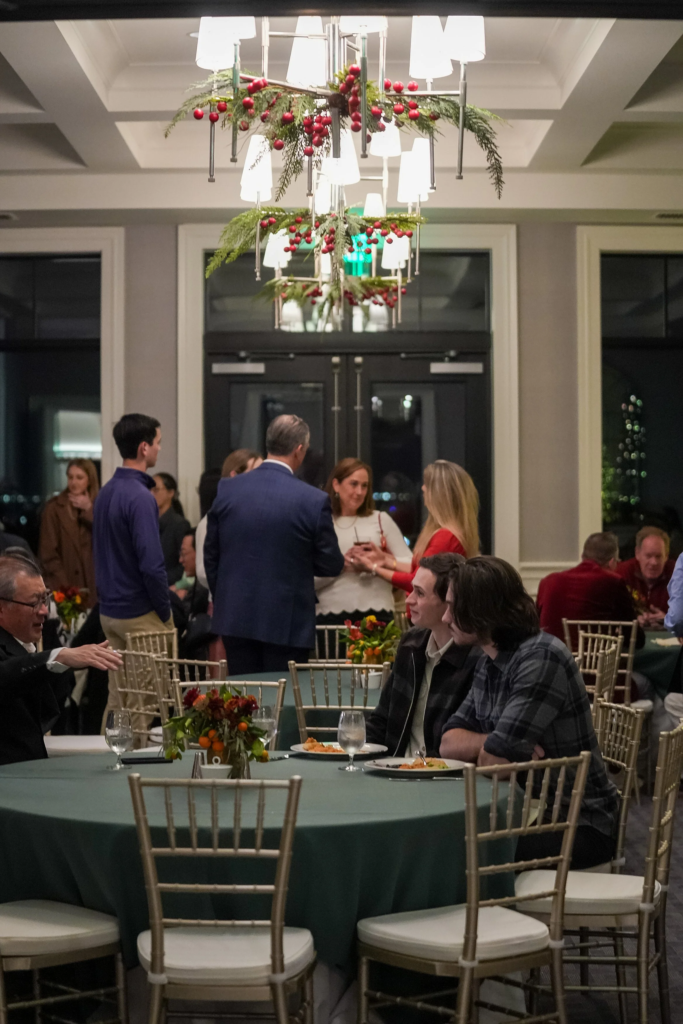 Group of people at a formal gathering seated around tables with green tablecloths, elegant chairs, floral centerpieces, and festive overhead decorations including red berries and greenery.
