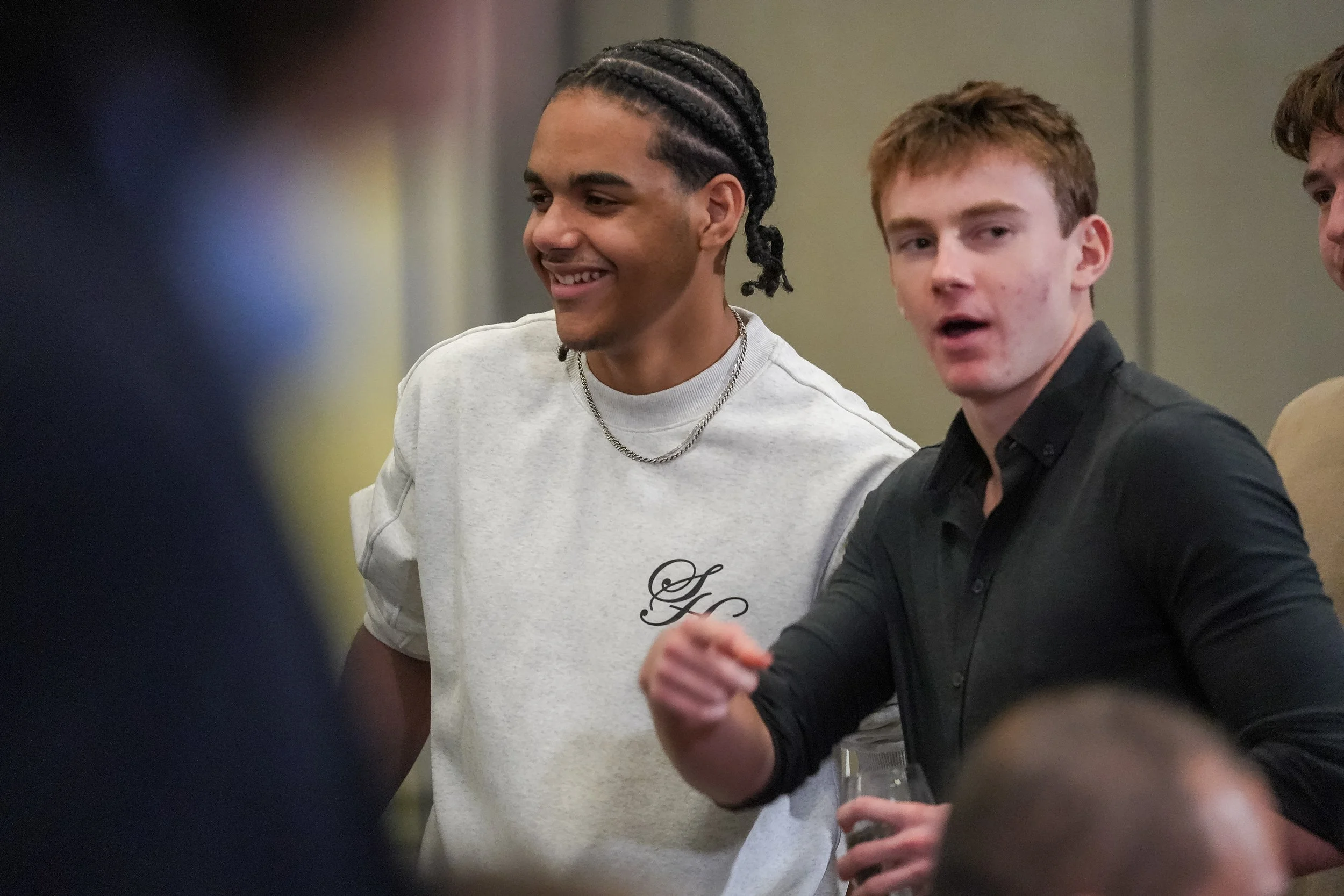 Two young men smiling and talking at an event; one has braided hair and wears a gray sweatshirt, the other has short hair and wears a black shirt.