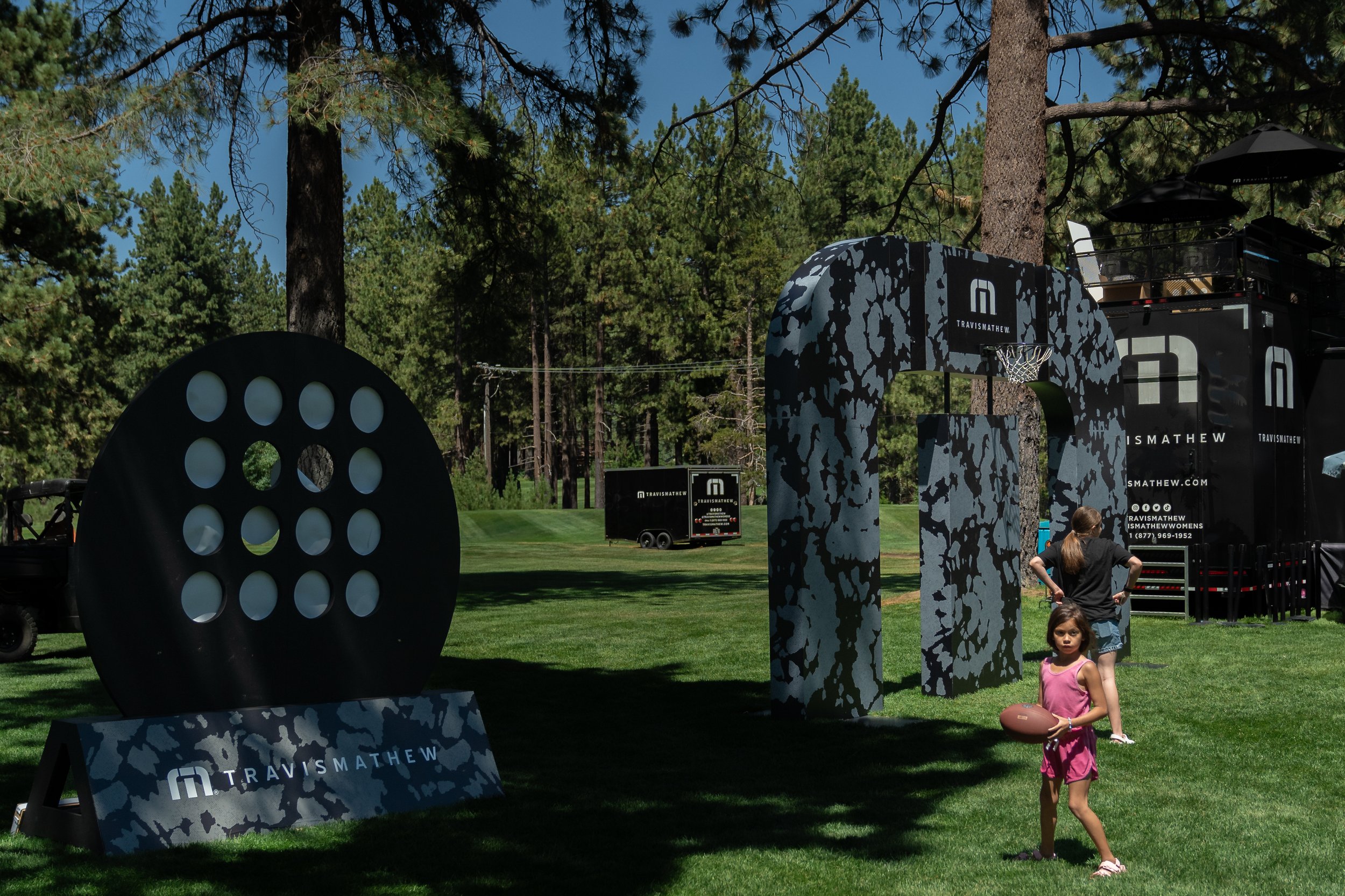 Outdoor event scene with TravisMathew branded structures and people playing on grassy area, surrounded by tall trees.