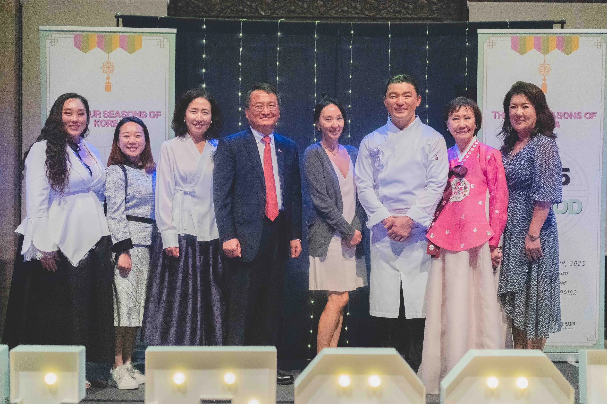 Group of nine people standing together on stage, dressed in formal, traditional, and professional attire, posing for a photo at an event with banners and decorative lights in the background.
