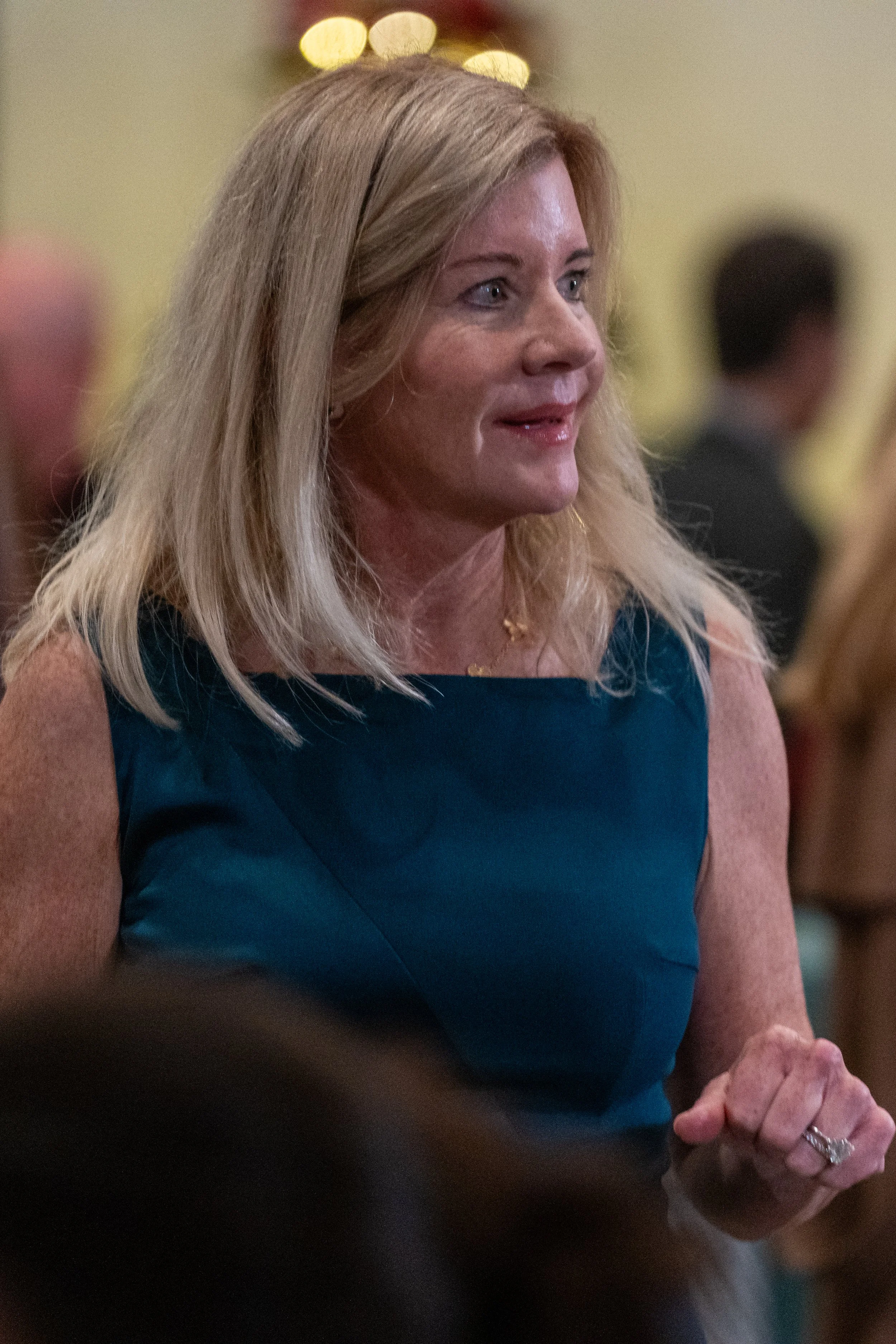 Blonde woman in teal dress at an event, surrounded by people in the background.