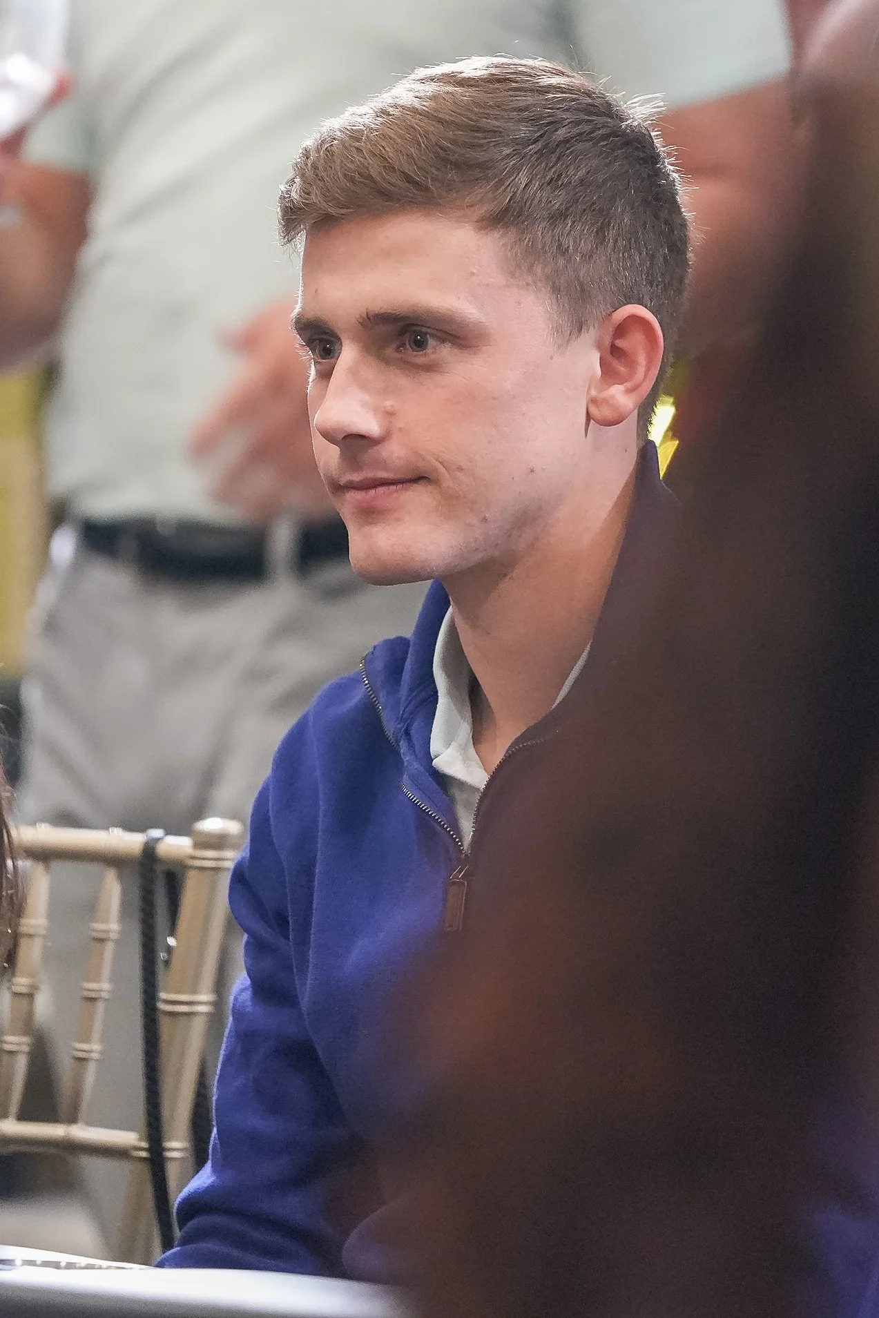 A man in a blue jacket sitting indoors, looking thoughtful.