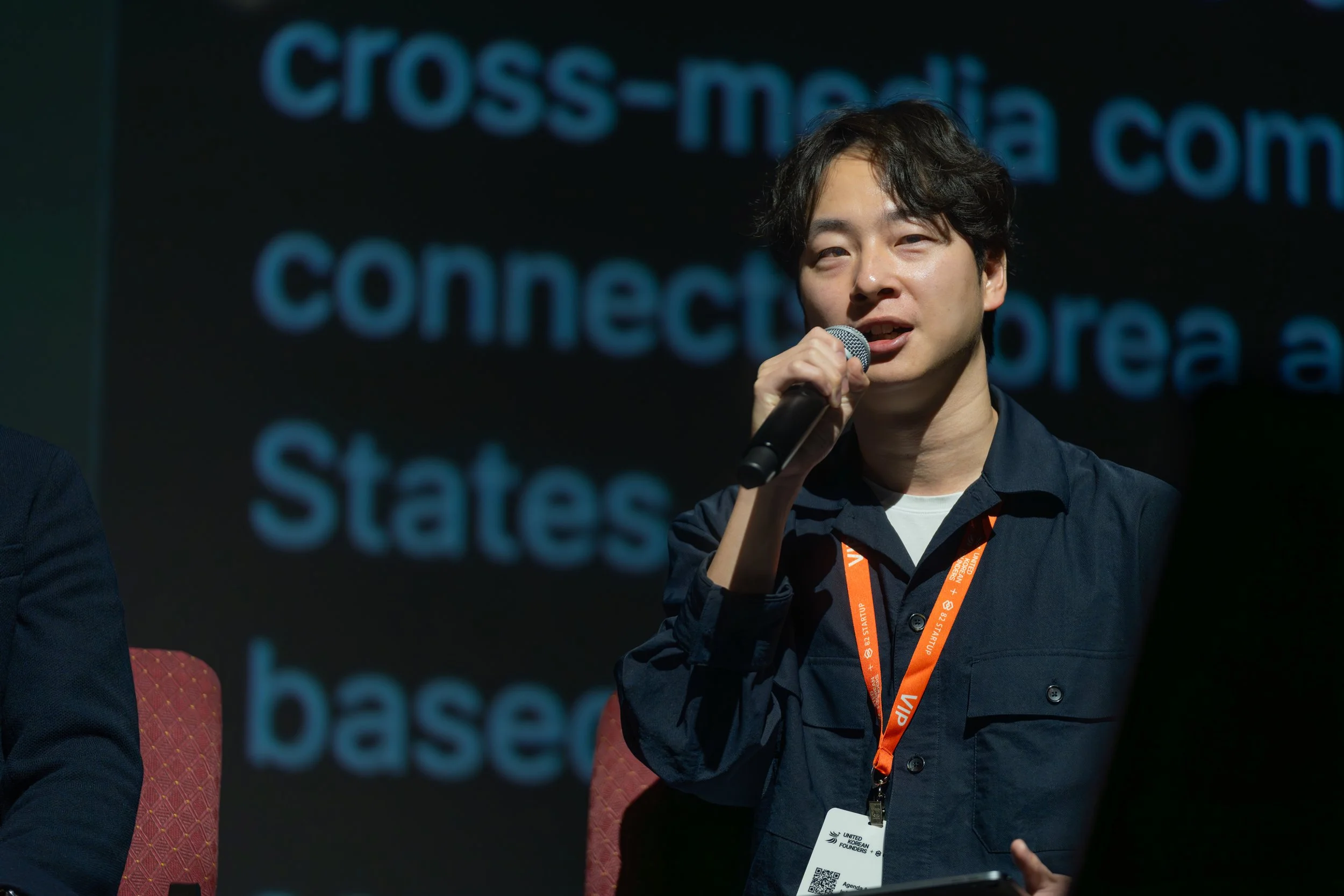 Person speaking into a microphone at an event with a lanyard and ID badge, background displays text.