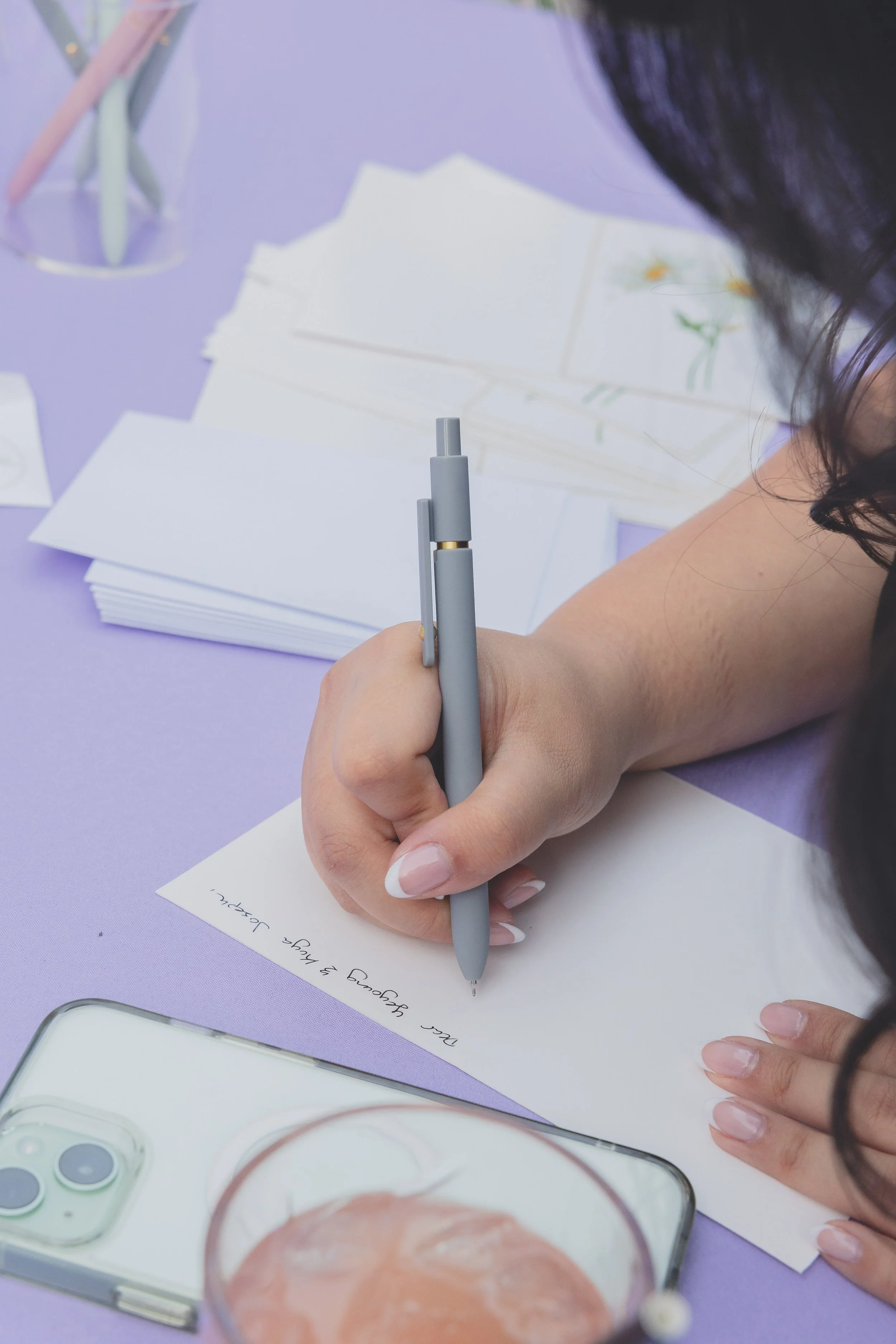 Person writing with a pen on white paper, surrounded by stacks of envelopes, with a smartphone and a drink on a purple table.
