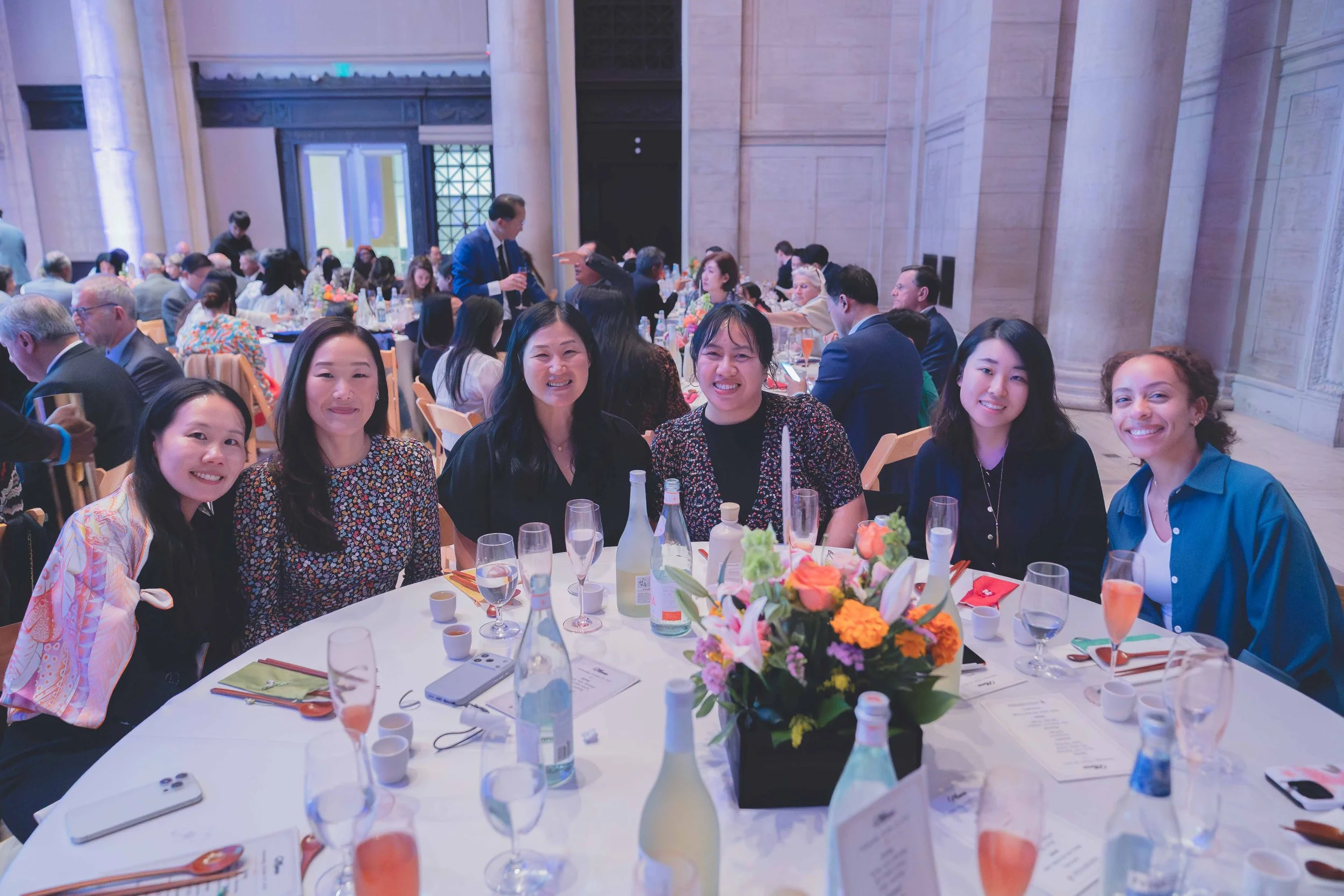 Group of six women sitting around a table at a formal event, smiling, with a floral centerpiece, glasses, bottles, and menus on the table, in a large decorated hall filled with other attendees.