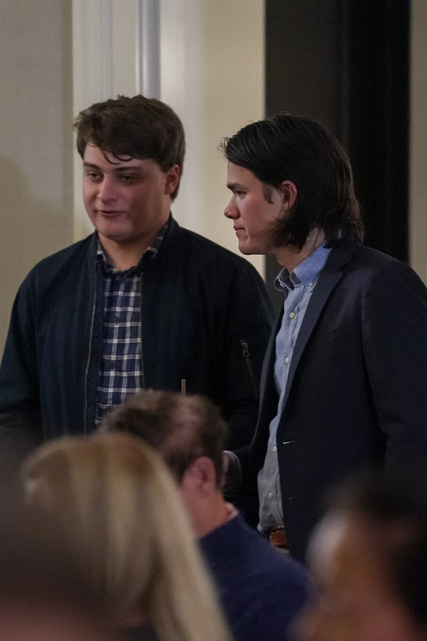 Two men standing indoors at an event, one wearing a plaid shirt and jacket, the other in a suit.