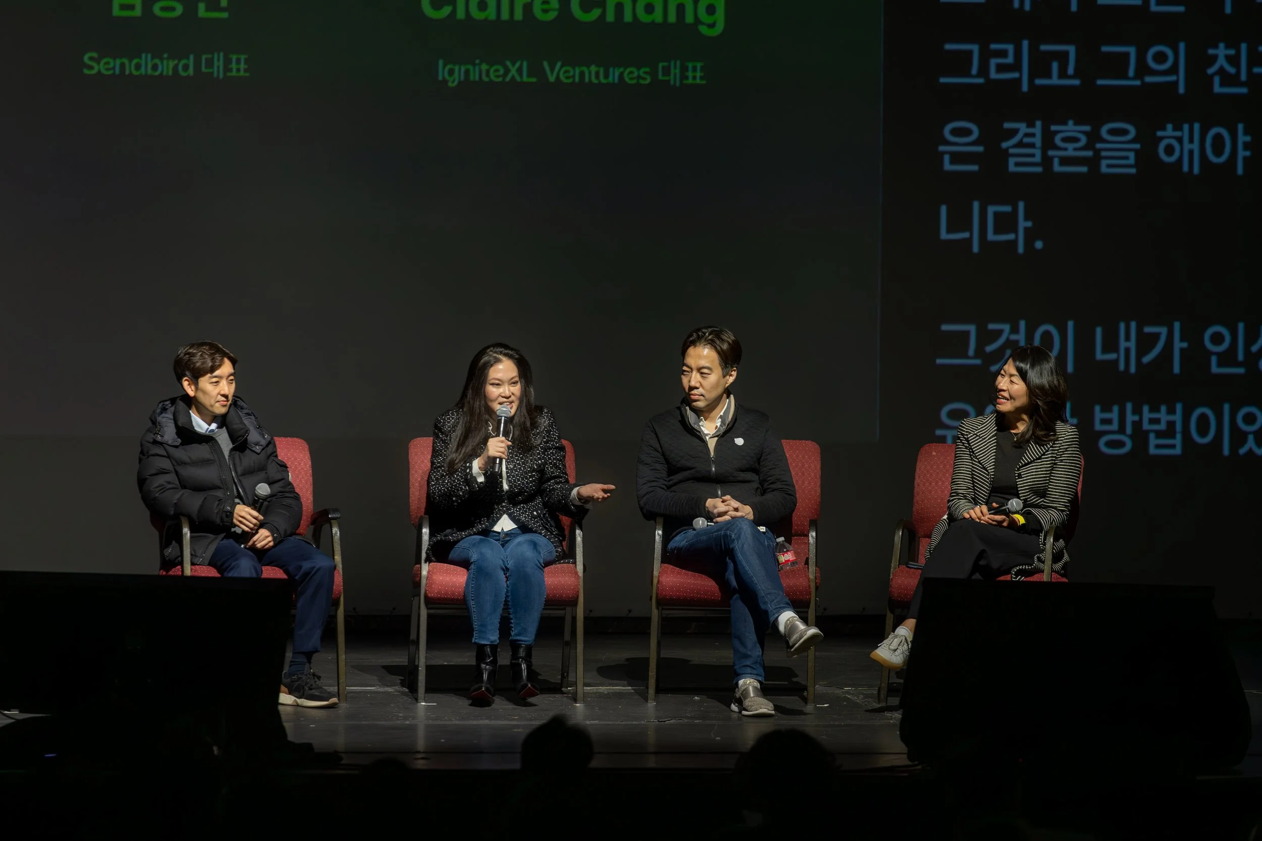 A panel of four speakers, two men and two women, seated on stage during a discussion. One woman is speaking into a microphone. The background features text in Korean and English.