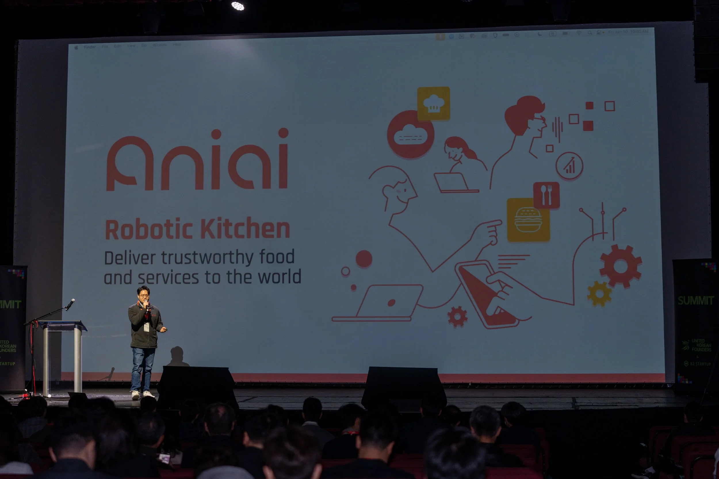 A speaker on stage presenting about 'Ani.ai Robotic Kitchen,' with a slide displaying technology and food-related graphics and the text 'Deliver trustworthy food and services to the world.' The audience is visibly present.