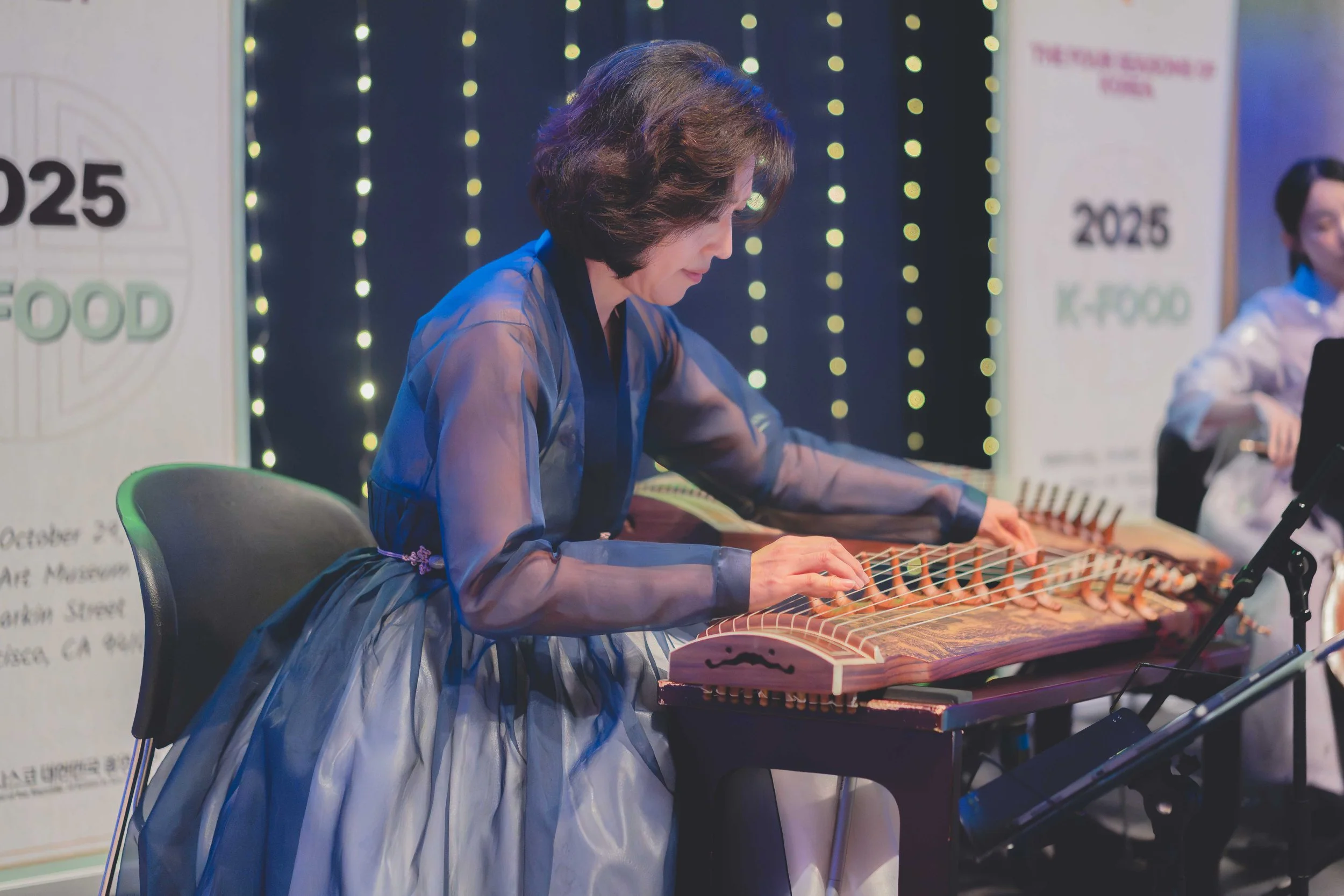A woman playing a gayageum, a traditional Korean string instrument, during a performance at an event with a festive backdrop of string lights and banners.