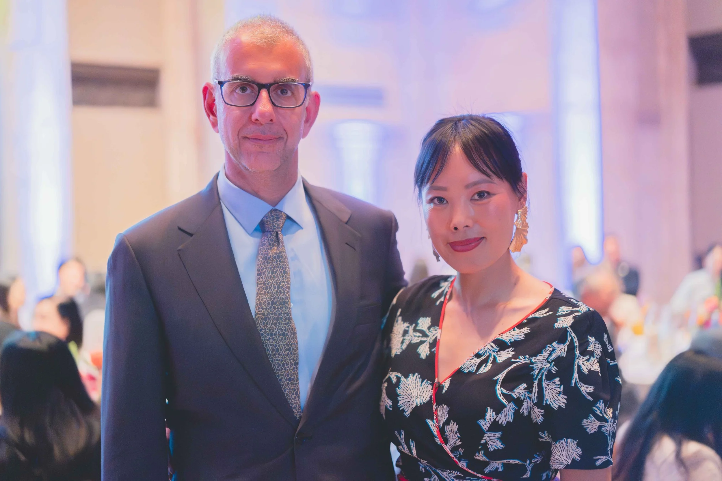 A man and a woman dressed in formal attire, posing together at an indoor event with blurred guests in the background.
