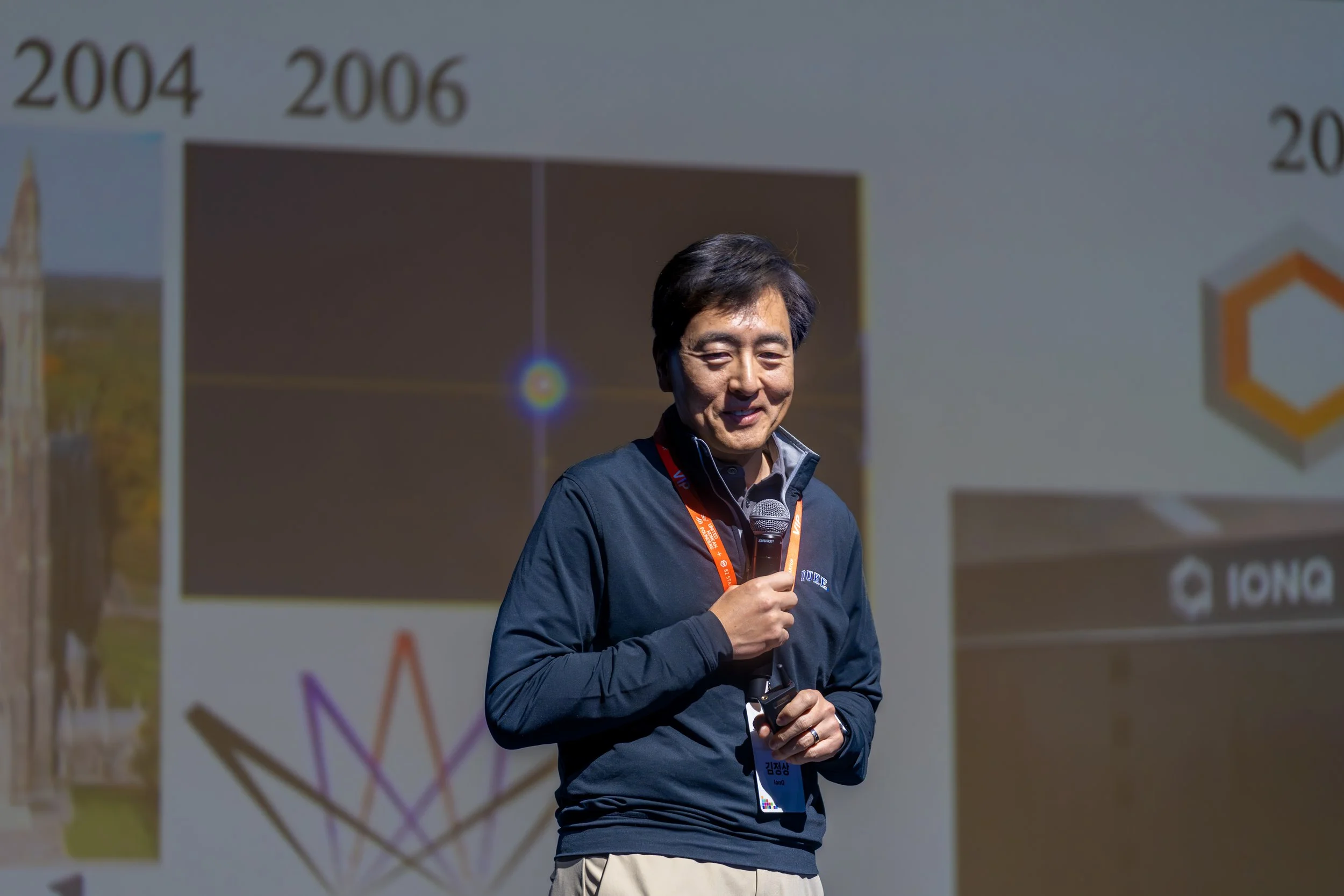 Person speaking on stage with a microphone, wearing a lanyard and dark jacket, with a presentation slide in the background.