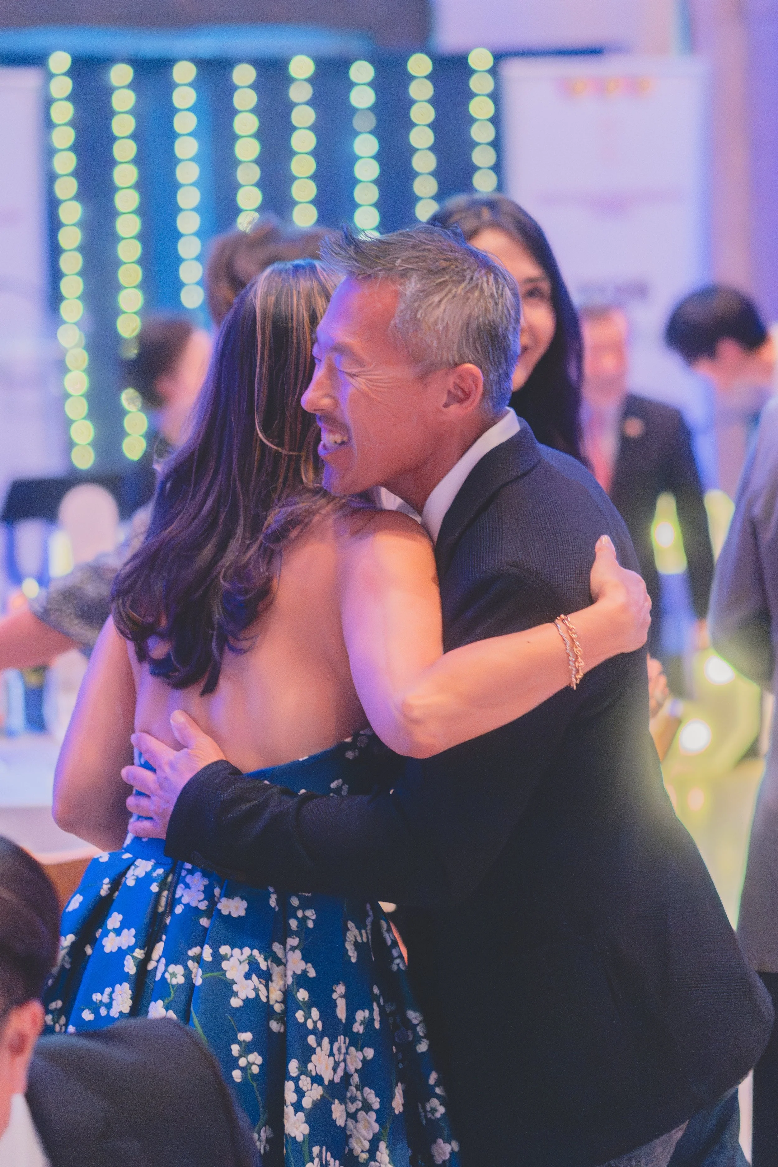 Two people hugging at a celebration with blurred background lights and other guests in formal attire.