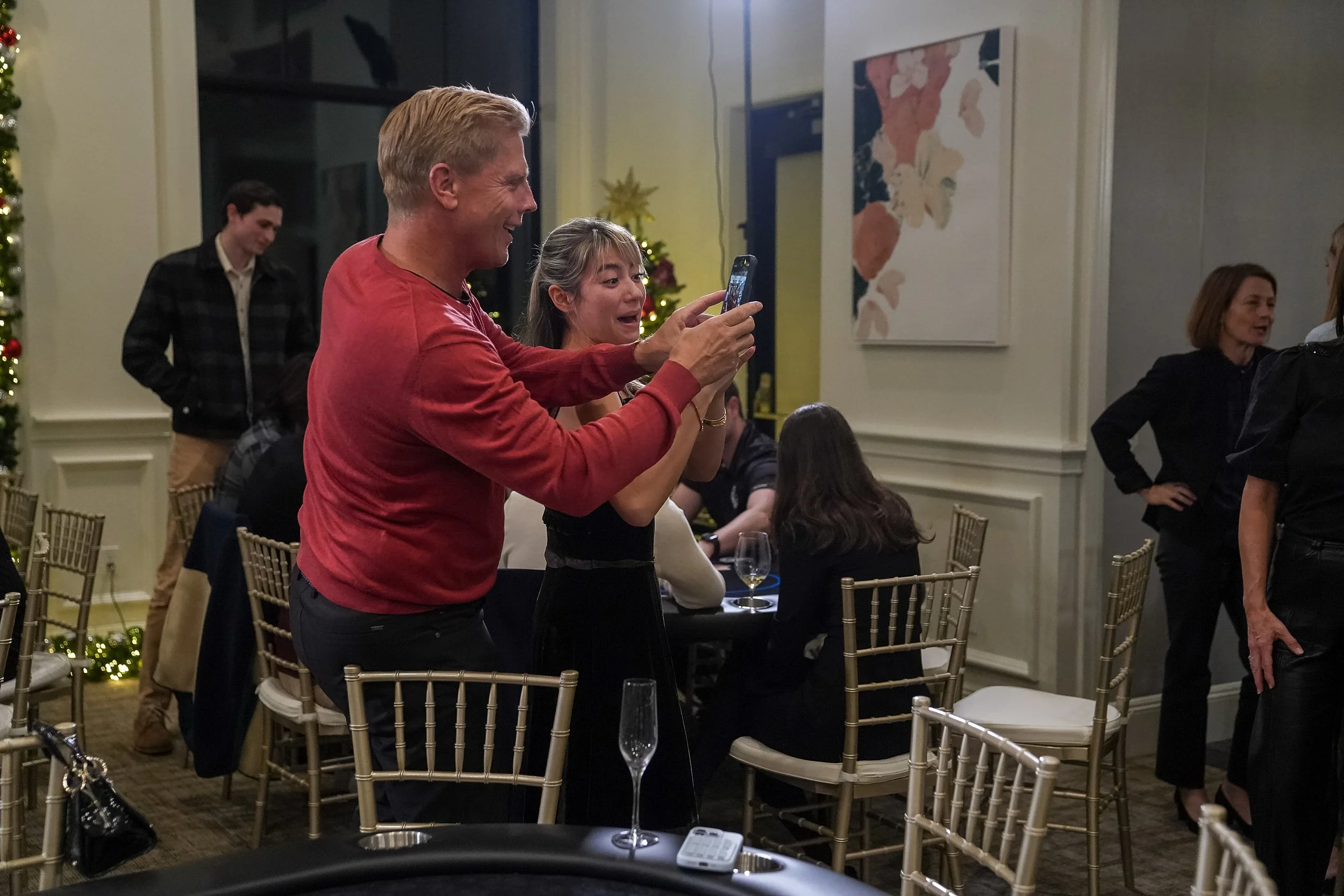 People at an indoor event taking a photo with a smartphone, surrounded by chairs and tables, with festive decorations in the background.