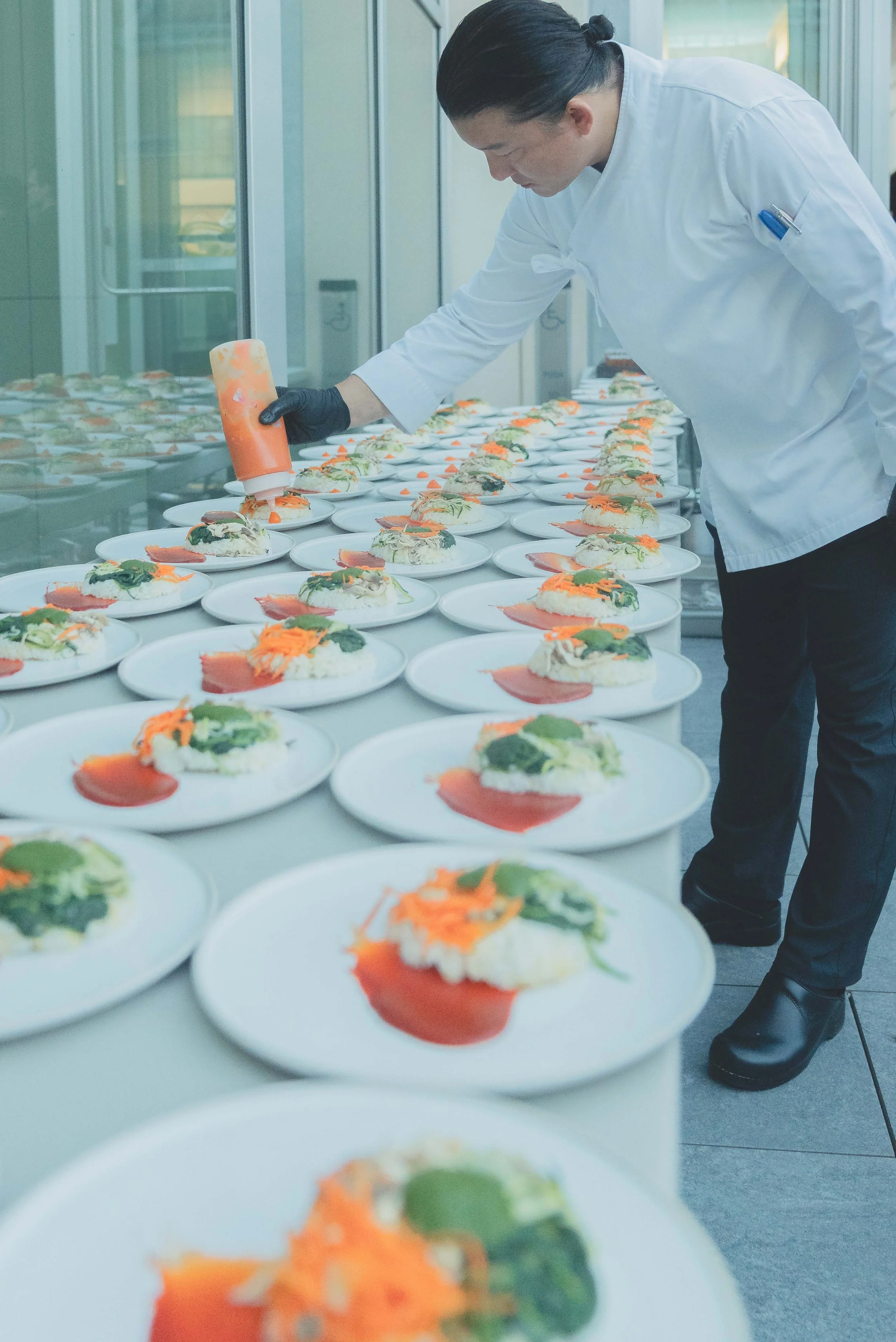 Chef in a white coat and black gloves decorating multiple plates of food with orange sauce in a professional kitchen.