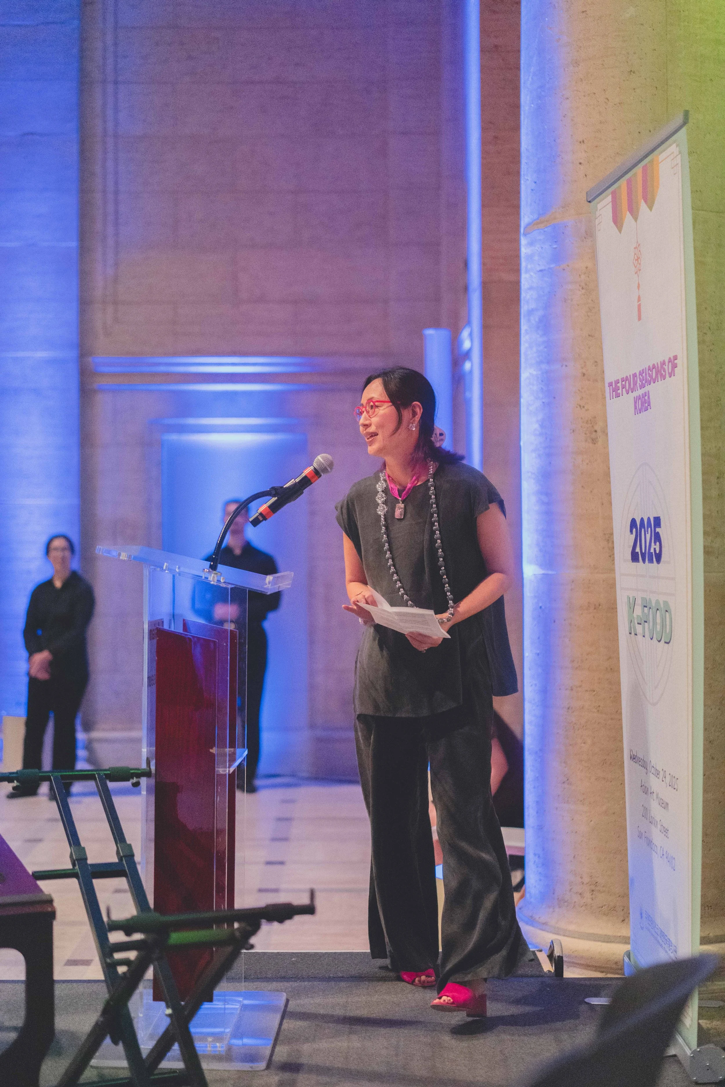 A woman with black hair and red glasses speaking at a podium during an indoor event, with a colorful banner on the side that reads 'The Four Seasons of Korea, 2050, K-Food' and a few people in the background.