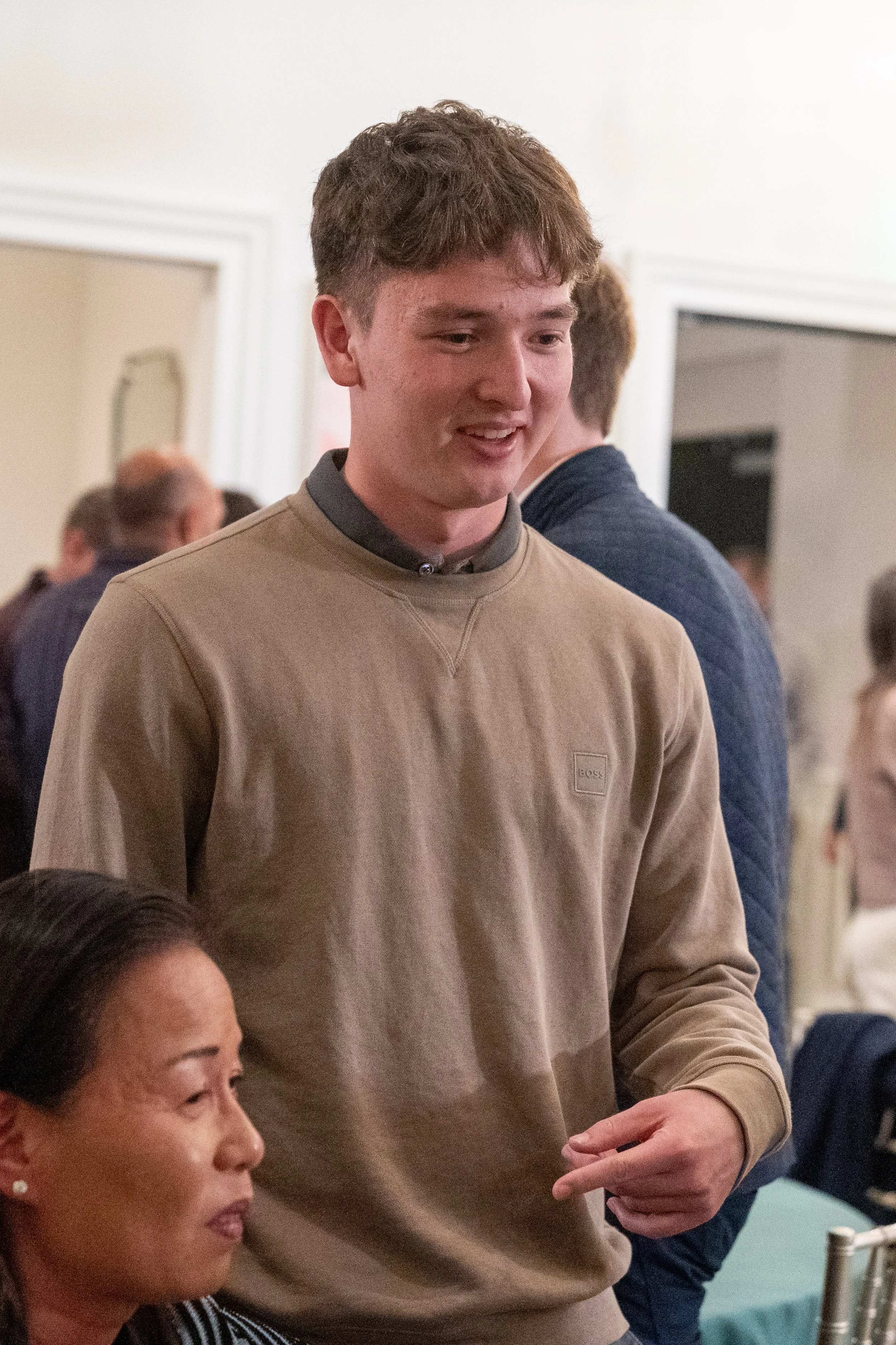 Young man in a casual tan sweater standing indoors, speaking to a seated woman in a lively setting.