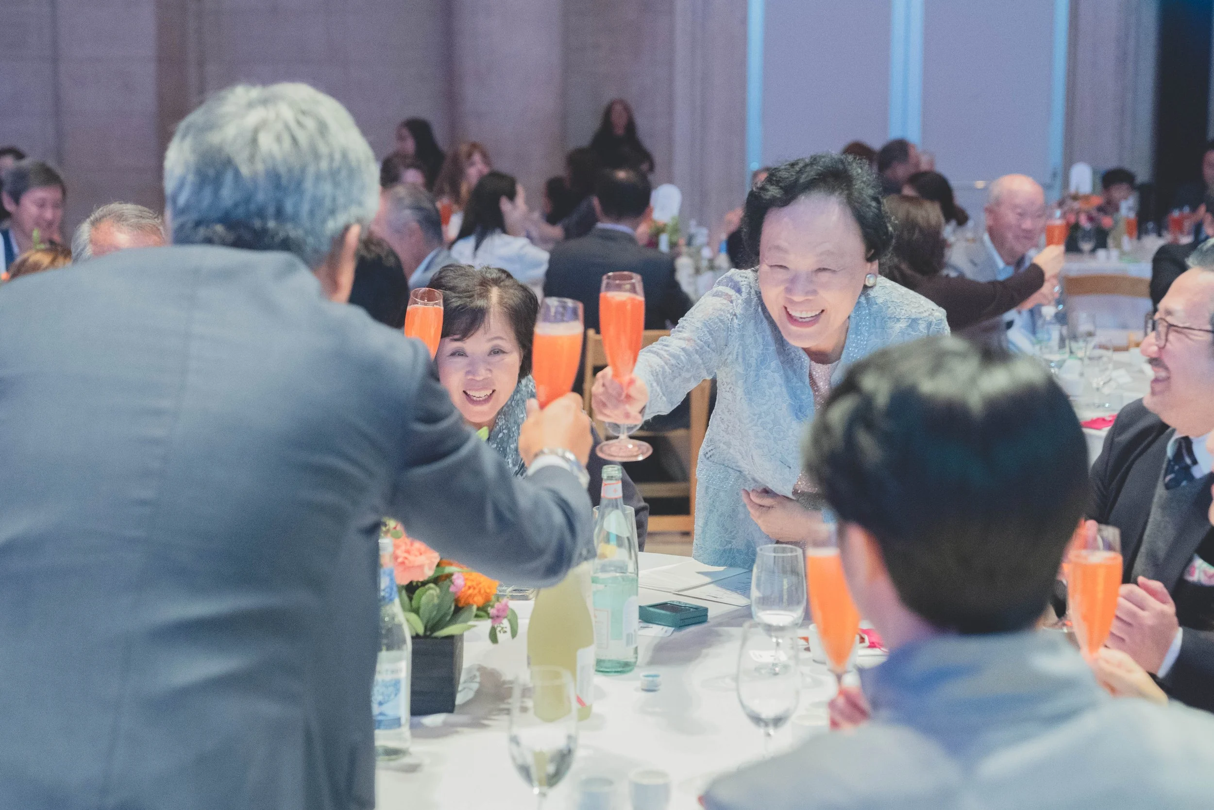 People at a formal event raising glasses of pink champagne, smiling and toasting, with a floral centerpiece on the table.