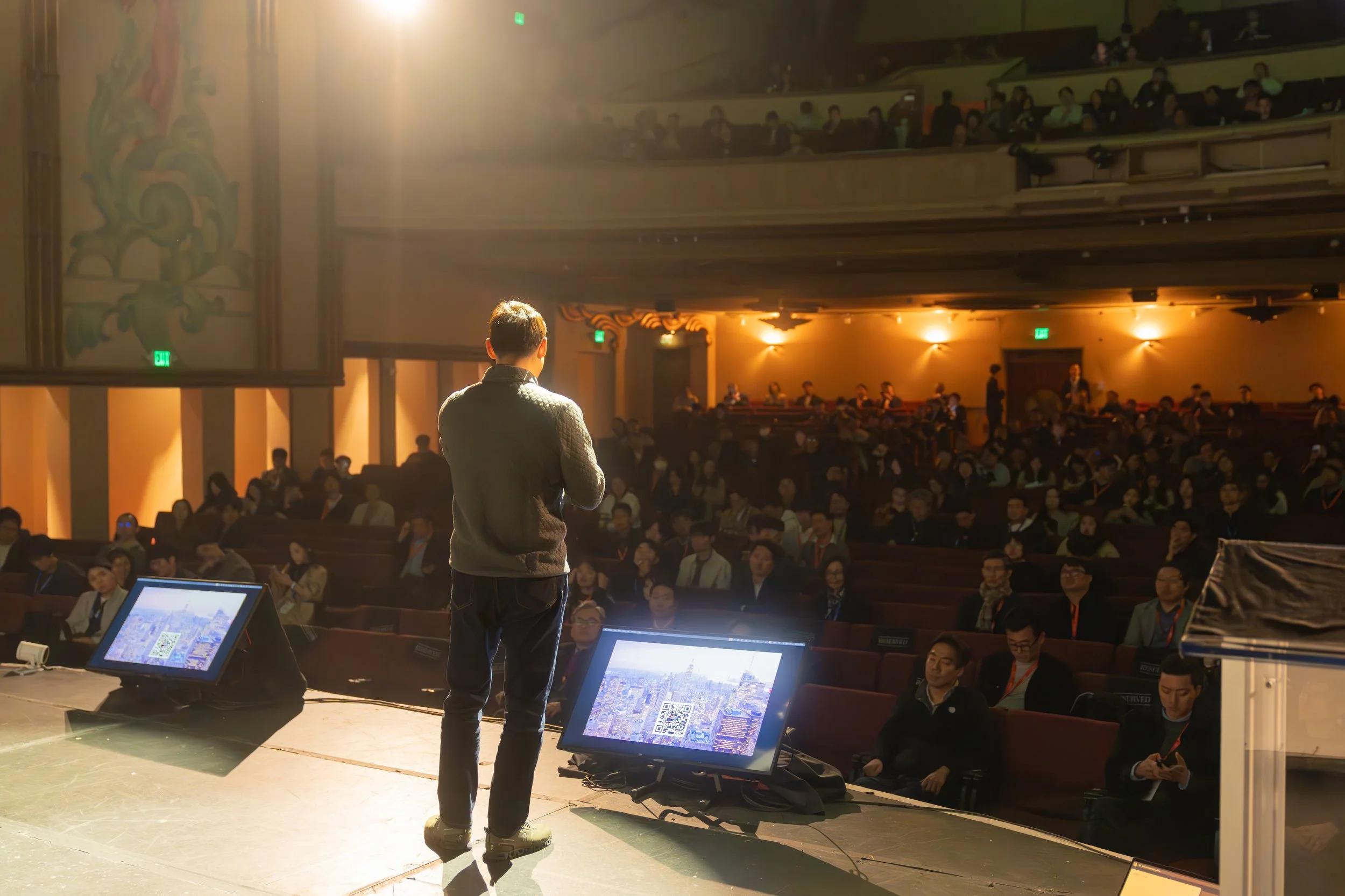 Speaker on a stage addressing an audience in a theater setting at a conference, with two screens displaying a cityscape and QR code.
