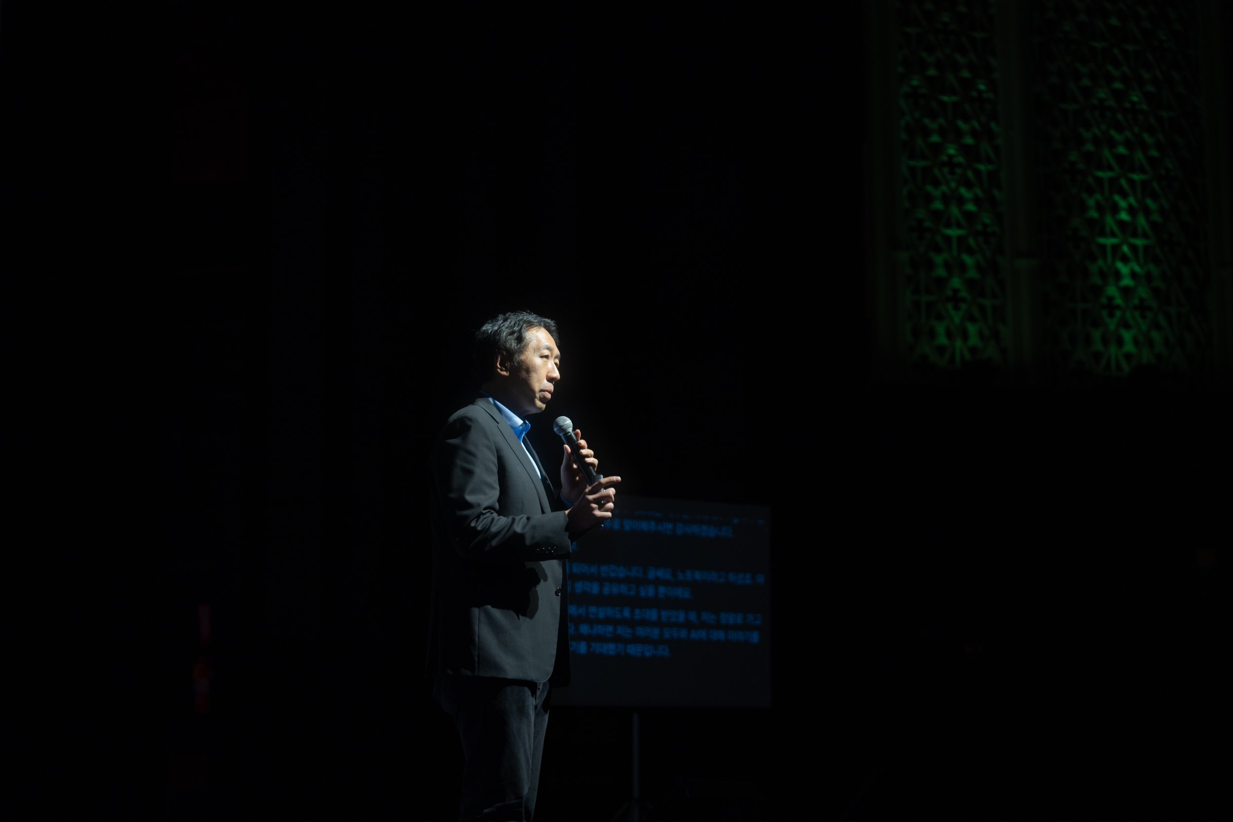 A man in a suit holding a microphone, speaking on stage with a prompter displaying text behind him. The background is dark with green decorative lighting.