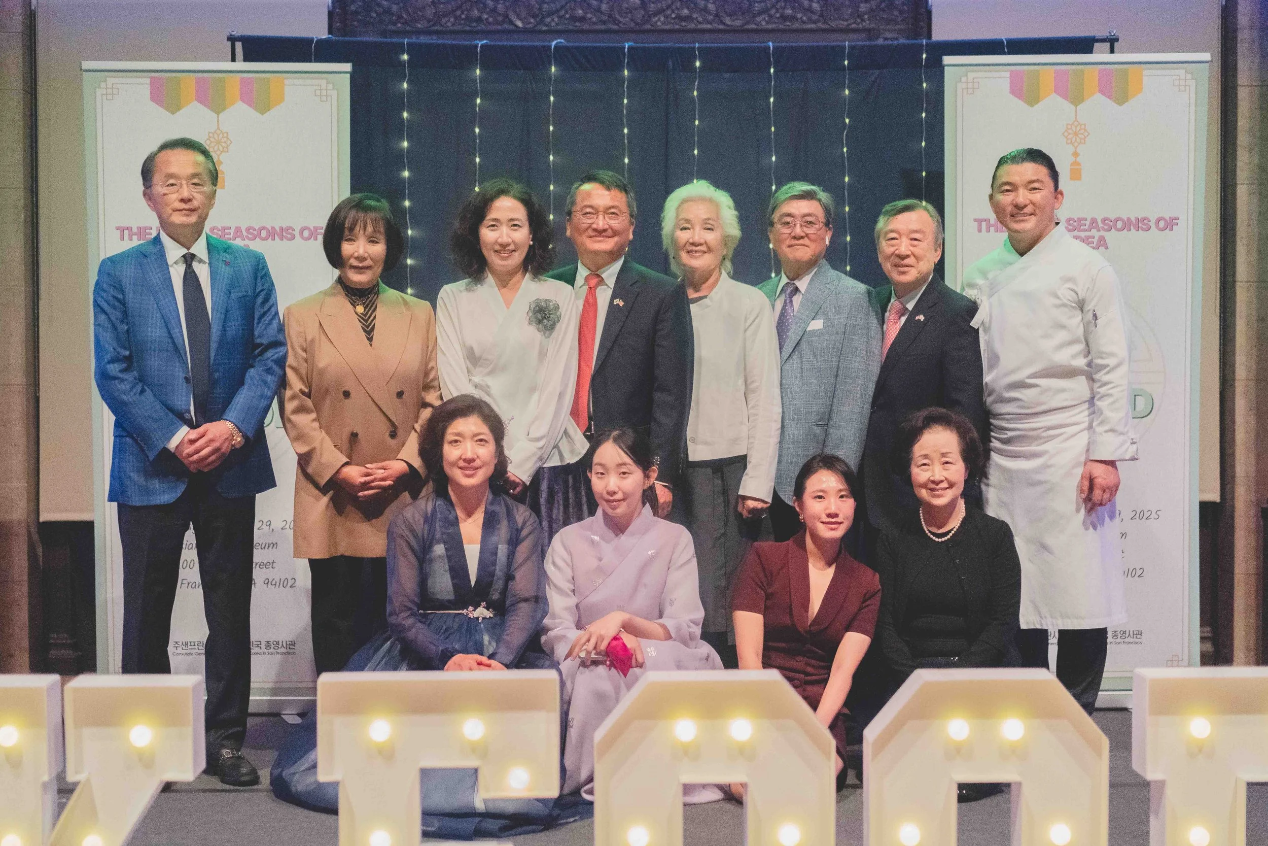 Group of people posing for a photo at an indoor event, with some in traditional Korean clothing and others in formal Western attire, standing in front of decorative banners and lights.