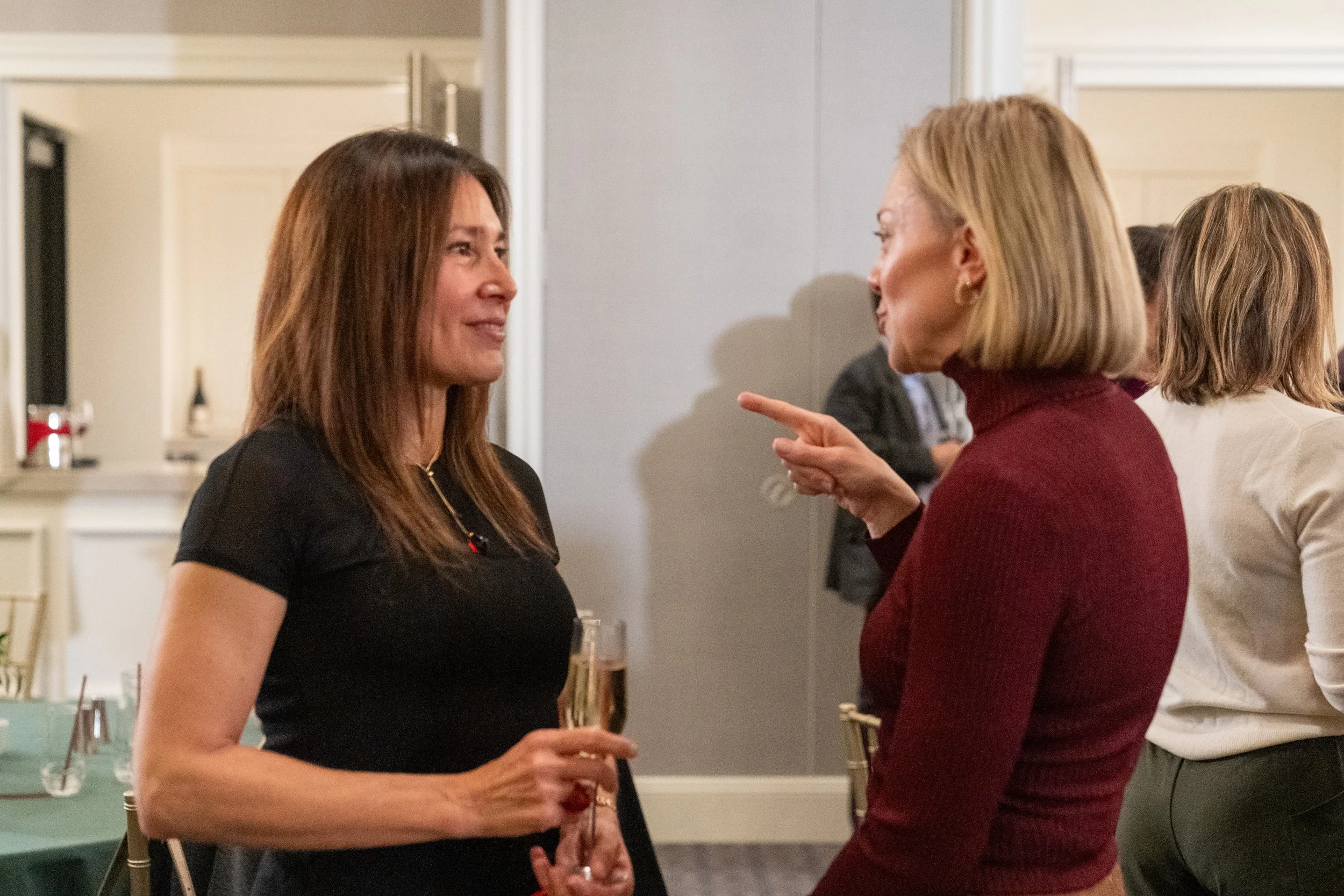 Two women having a conversation, one holding a glass, indoors at a social event.