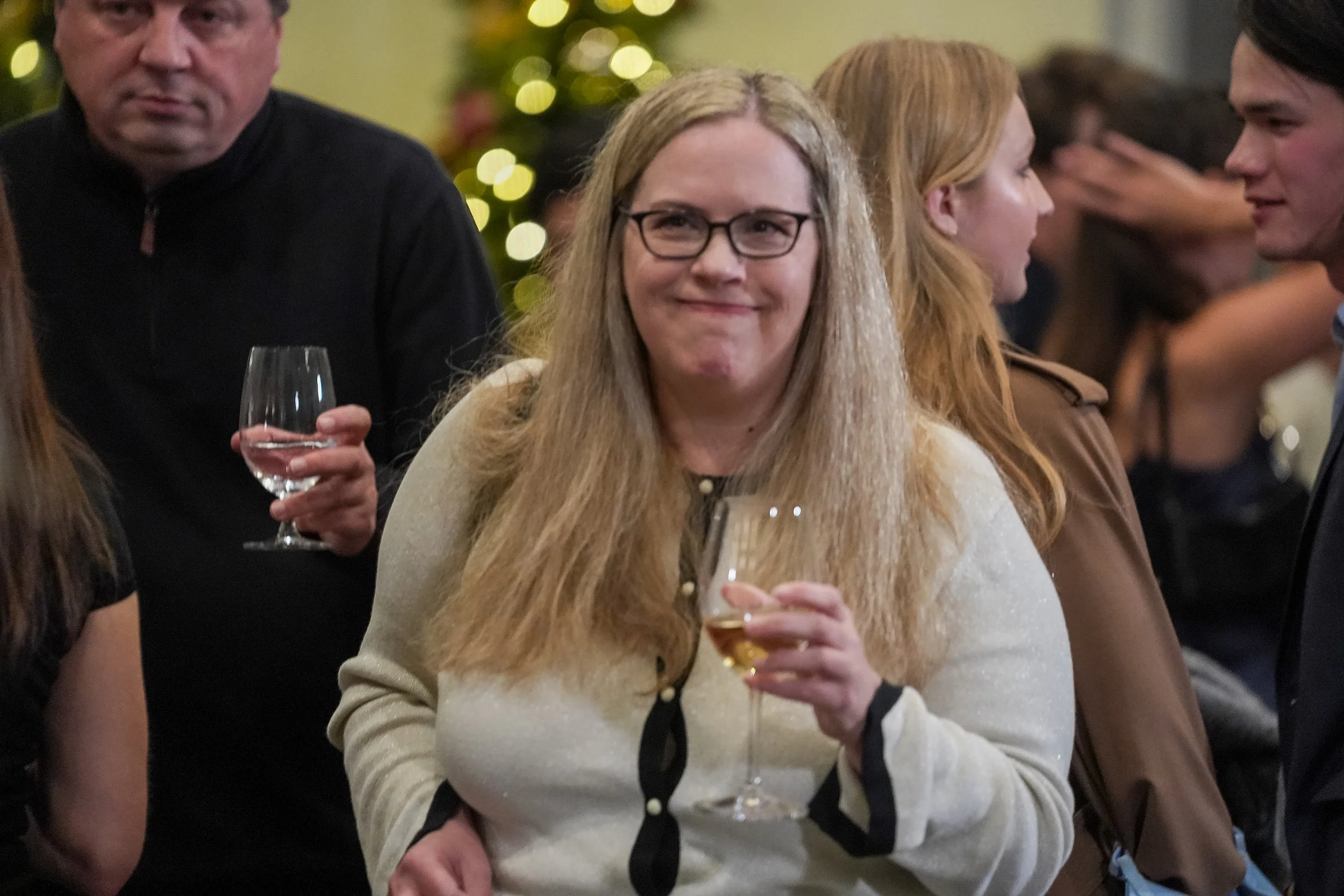 People socializing at an event, a woman in the foreground holding a wine glass, others in the background.