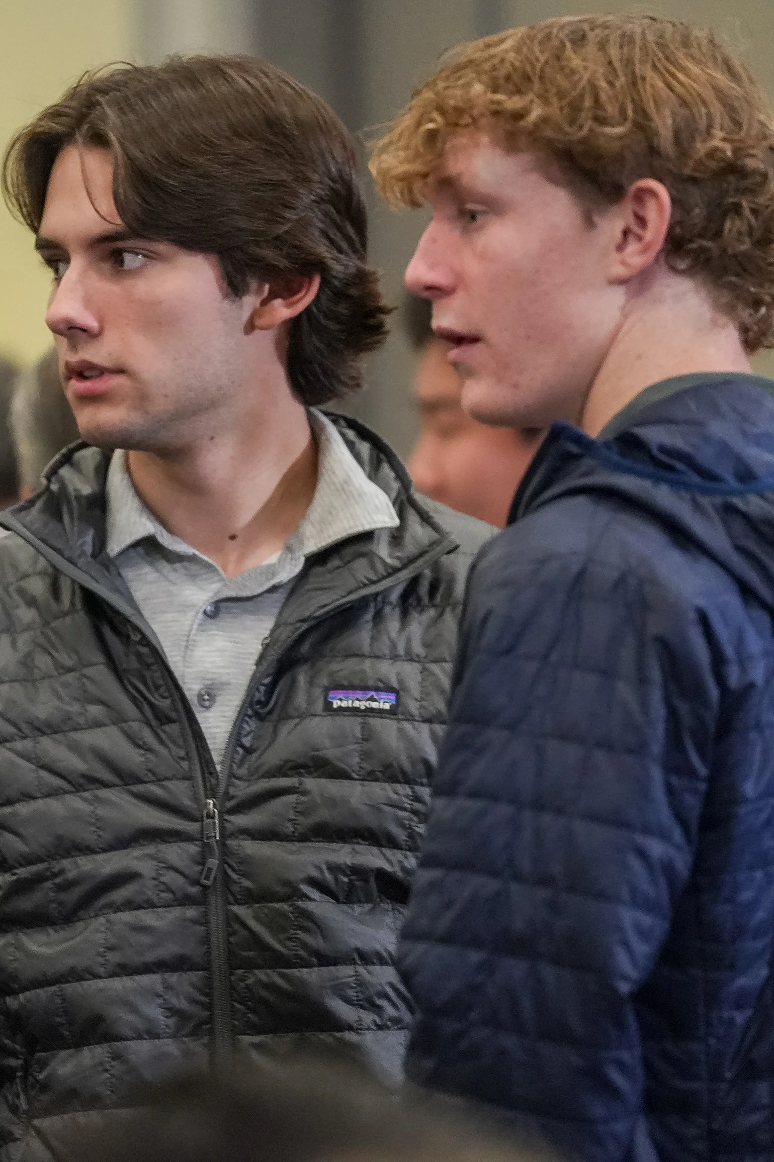Two young men wearing quilted jackets, talking indoors.