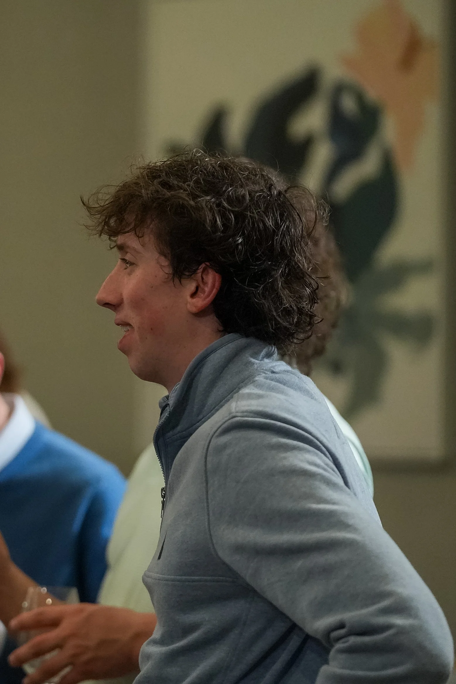 A person in a light blue jacket with curly hair is standing in profile view in a room with a blurred abstract painting in the background.